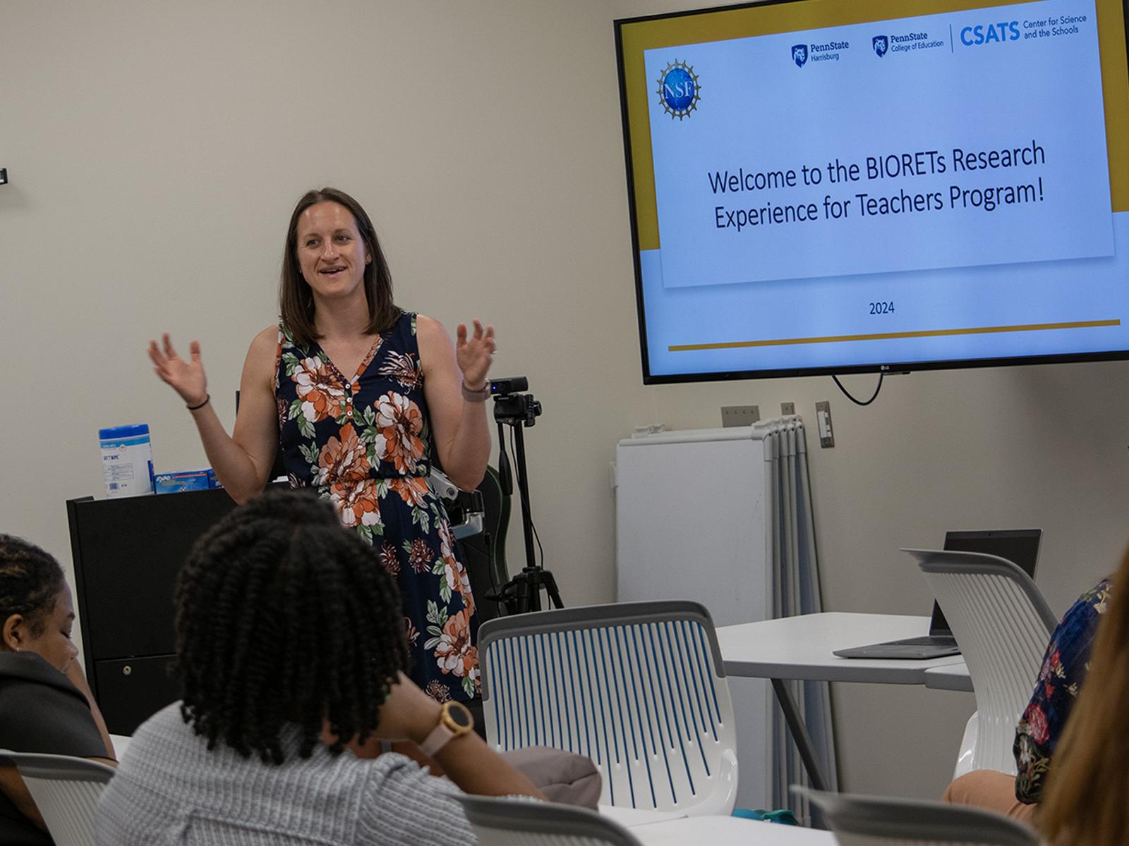 A woman stands in a classroom next to a screen that says "Welcome to the Biorets Research Experience for Teachers Program"