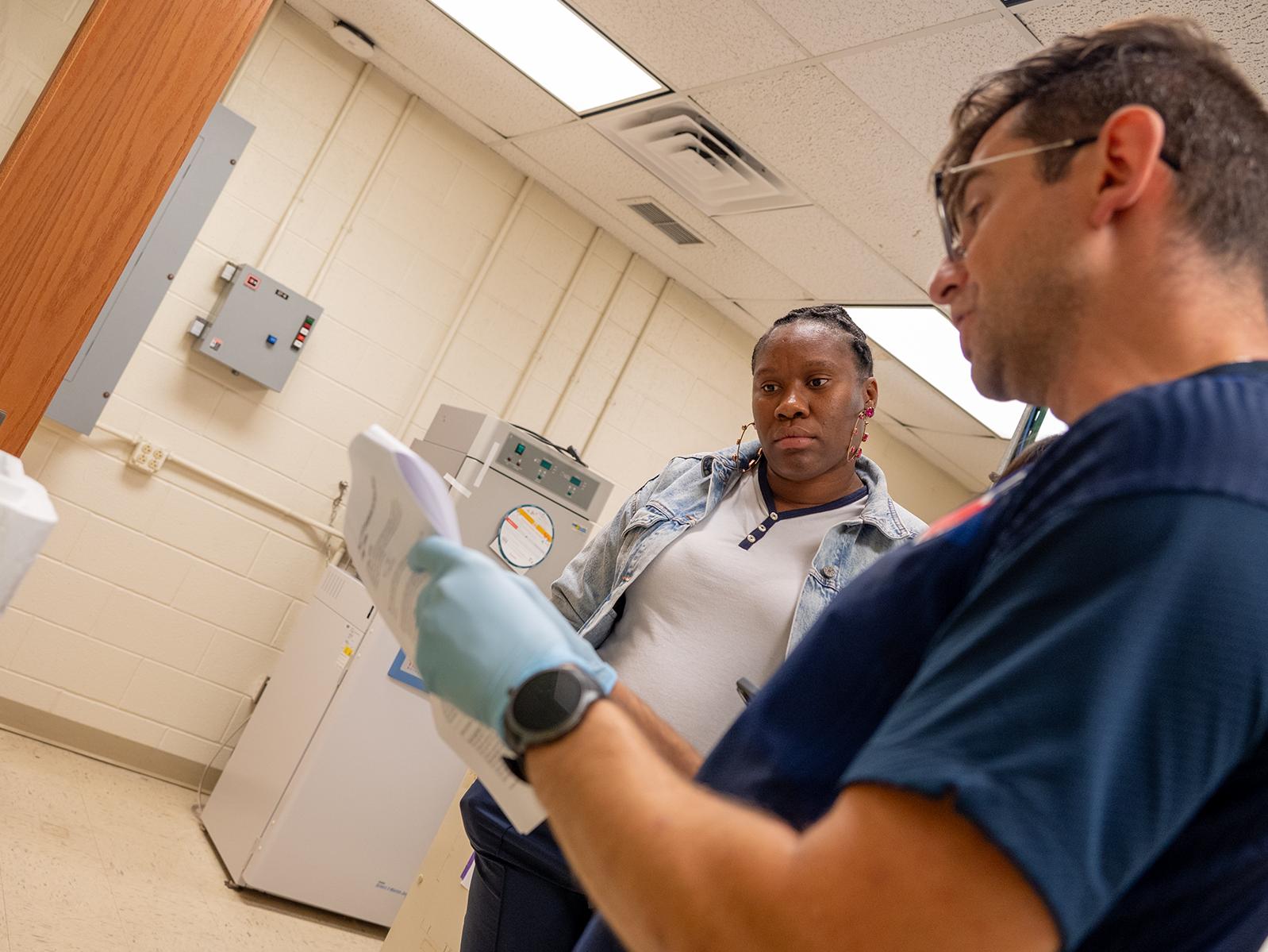 Two people stand in a lab, looking at a paper with instructions