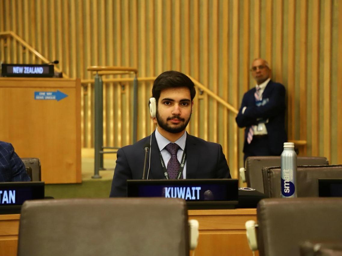 A student wearing a suit and headphones sits with a sign reading "Kuwait" in front of him