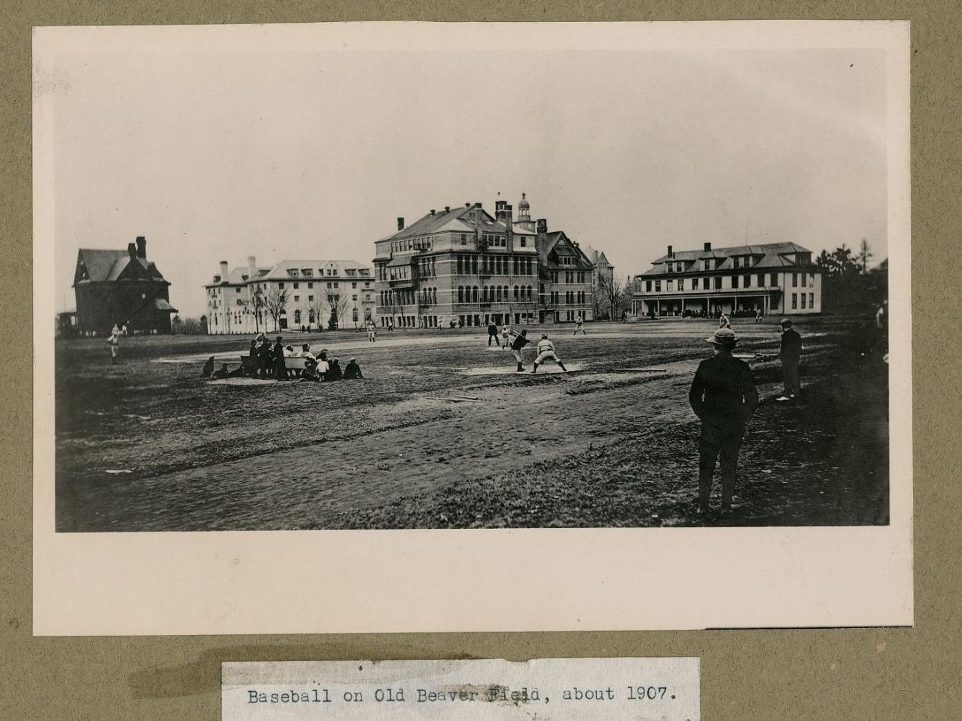 A picture of a baseball game c. 1907, taken from behind home plate. Four campus buildings can be seen in the background.