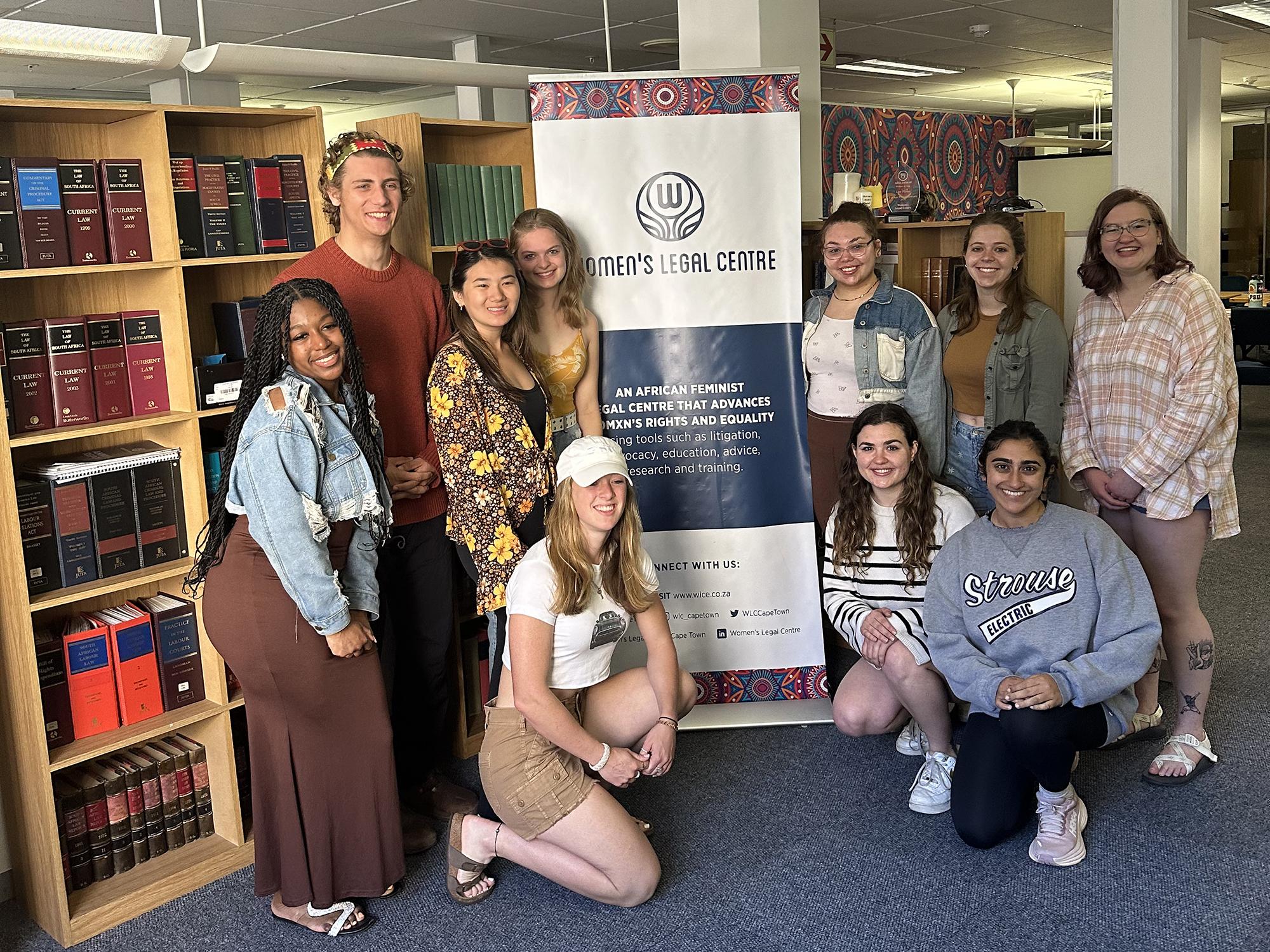 Students stand and kneel around a retractable banner reading Women’s Legal Centre in front of bookshelves.