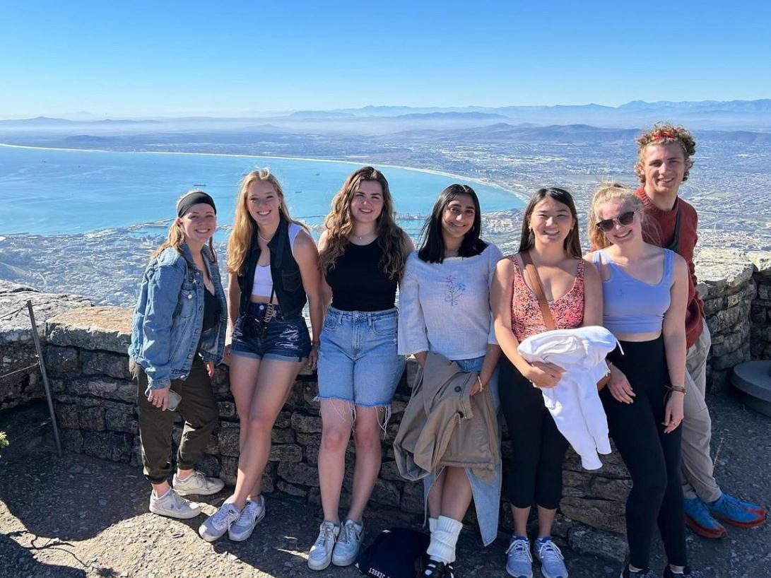 Seven students lean against a half wall of rocks on a ridge overlooking Cape Town, South Africa.