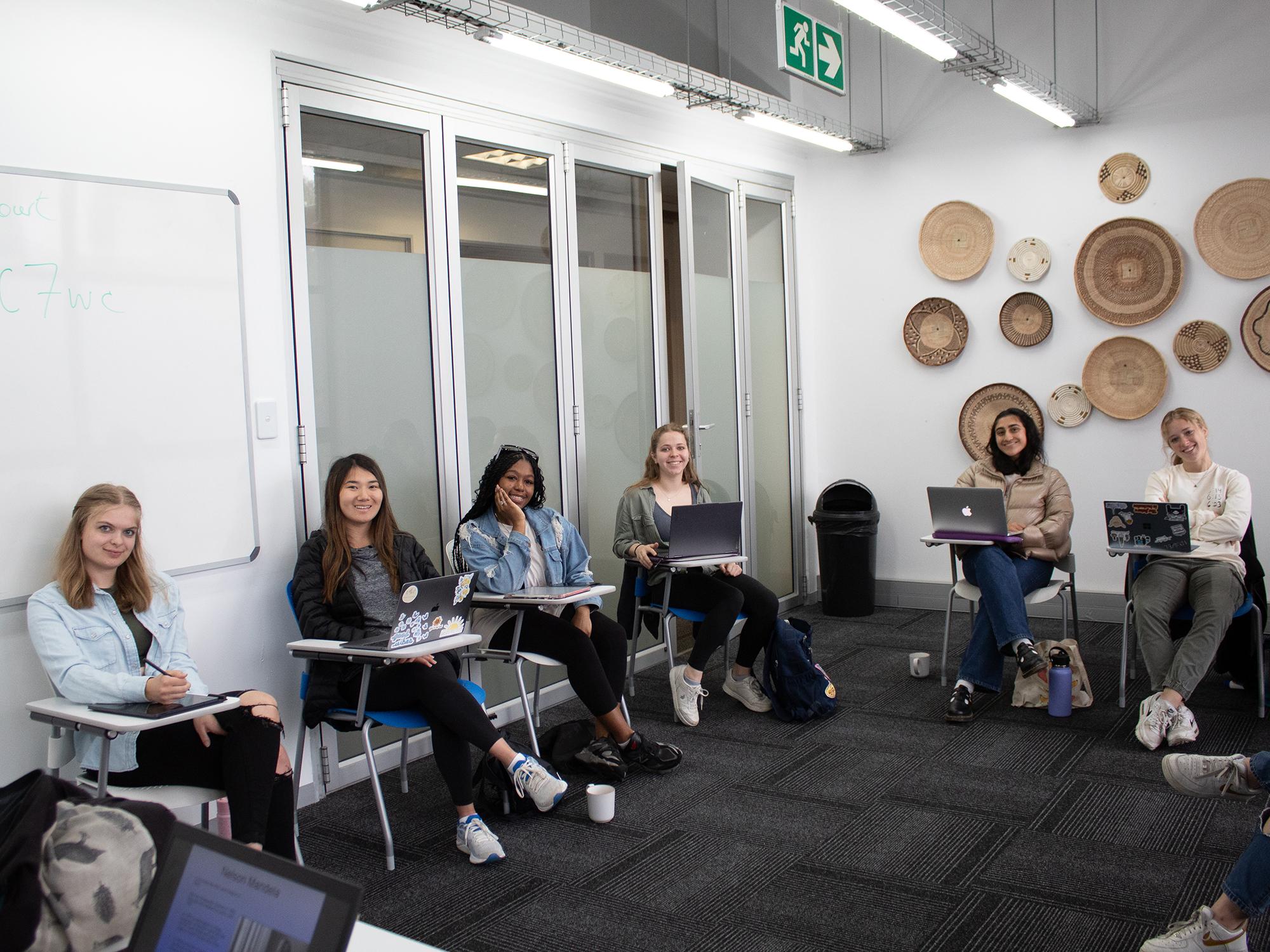 Six students sit in a semicircle of desks in their classroom.