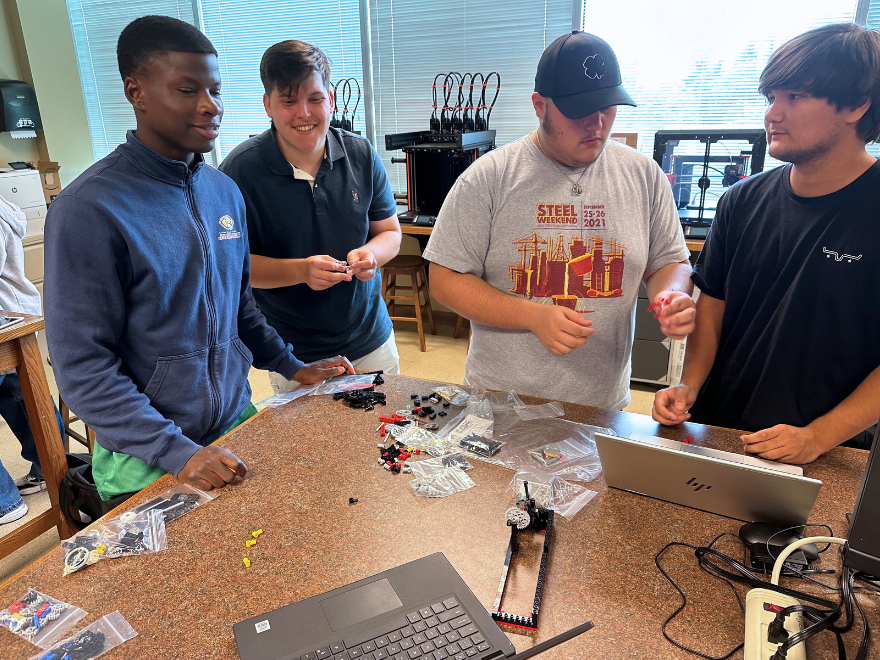 male college students collaborate on an engineering project at a long desk in a classroom