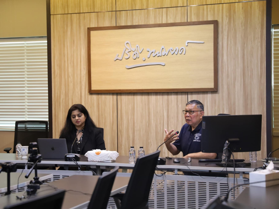 A man speaks to a group of people while sitting next to a woman behind a long desk
