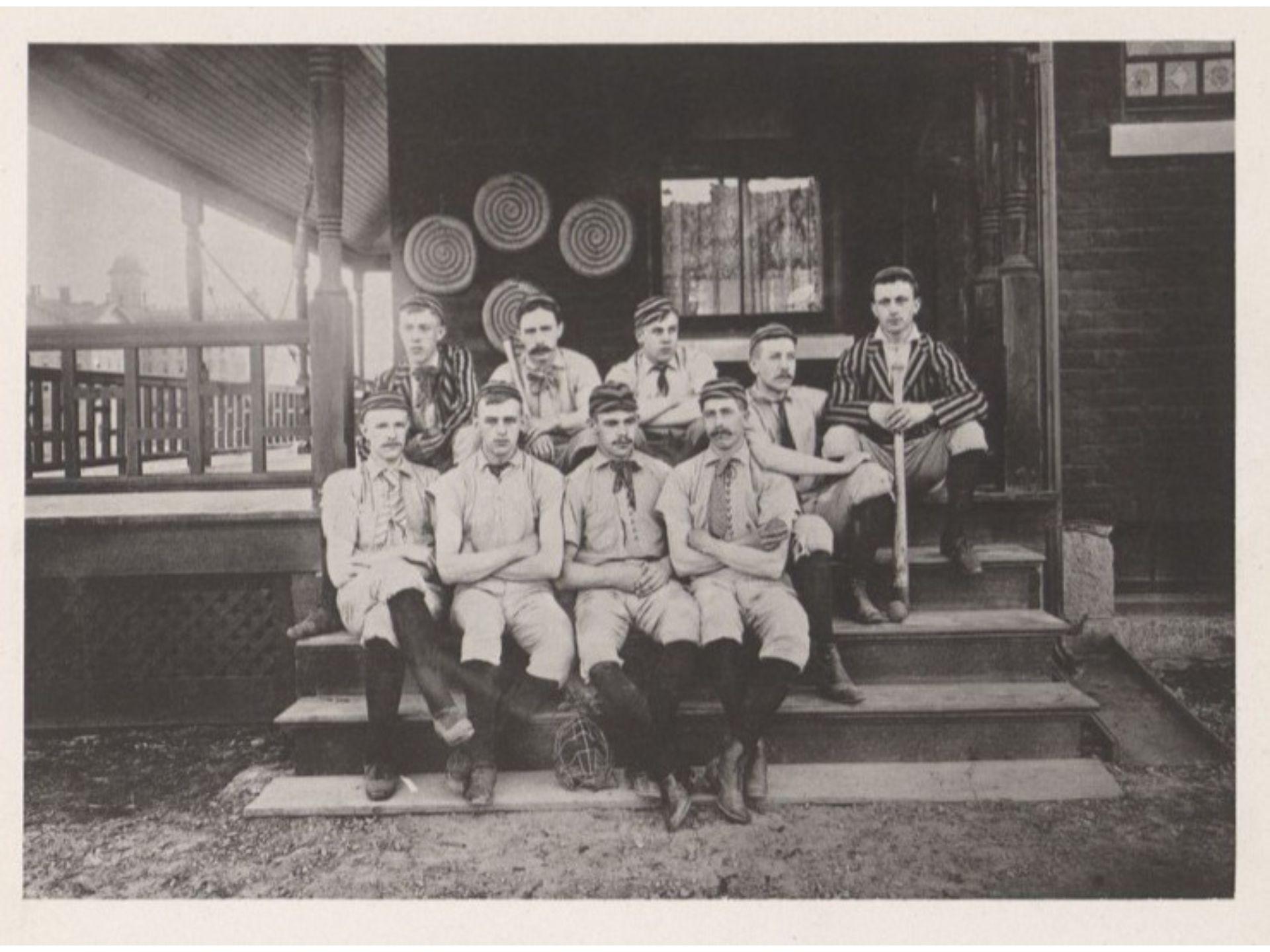 Nine baseball players sit on steps of a building in 1889. Two are wearing striped blazers and one is holding a baseball bat.