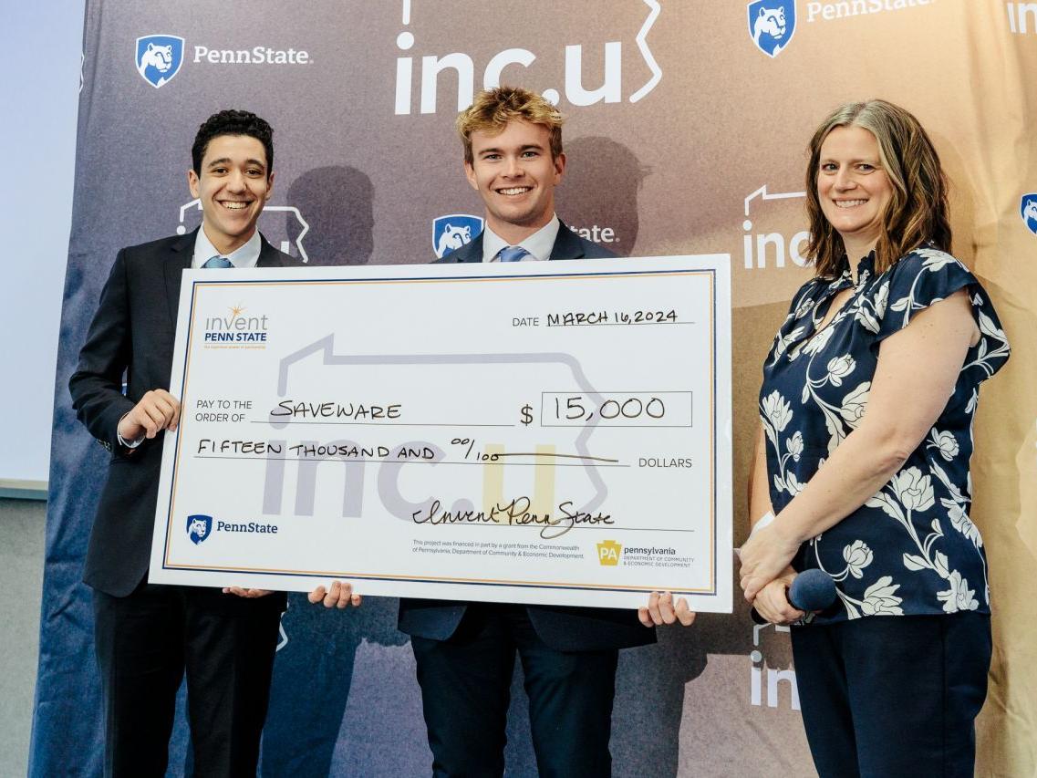 Two students hold a large check after winning a competition, with a woman presenting the check