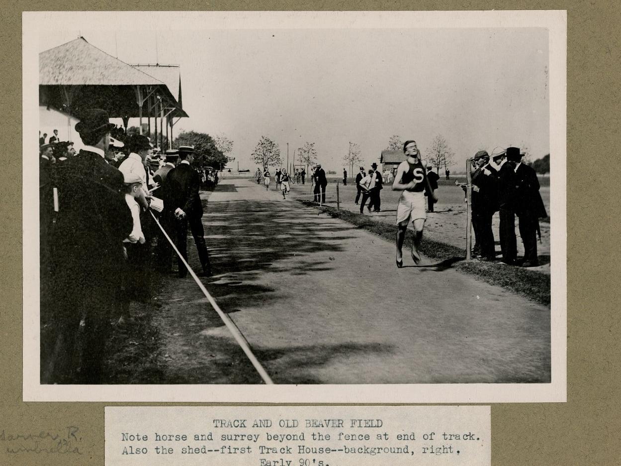 A runner wearing an S on his uniform funs past a crowd on a track in the 1890s