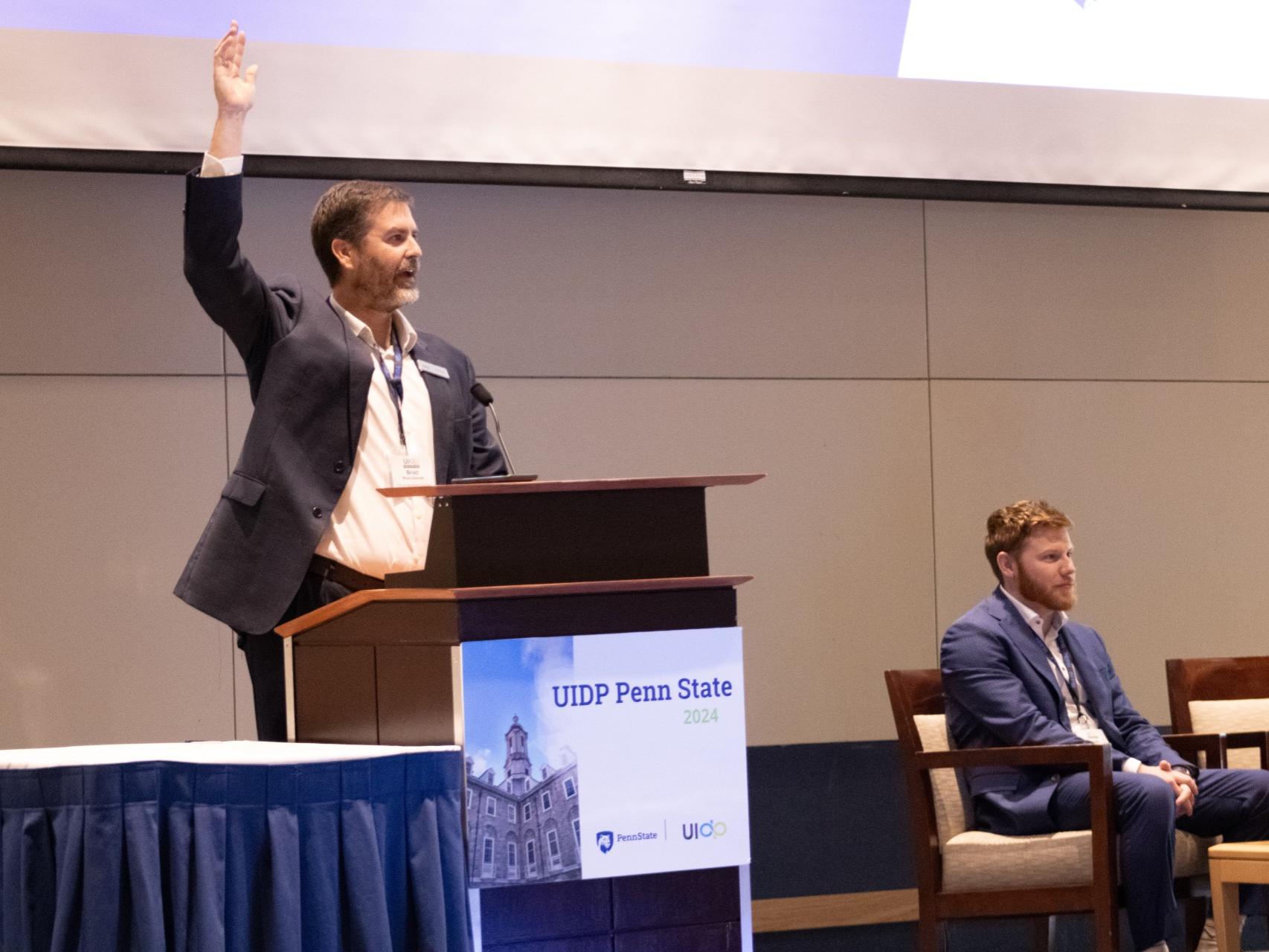 Person standing at a podium with their right hand raised, someone is sitting in a chair on the stage next to the podium.