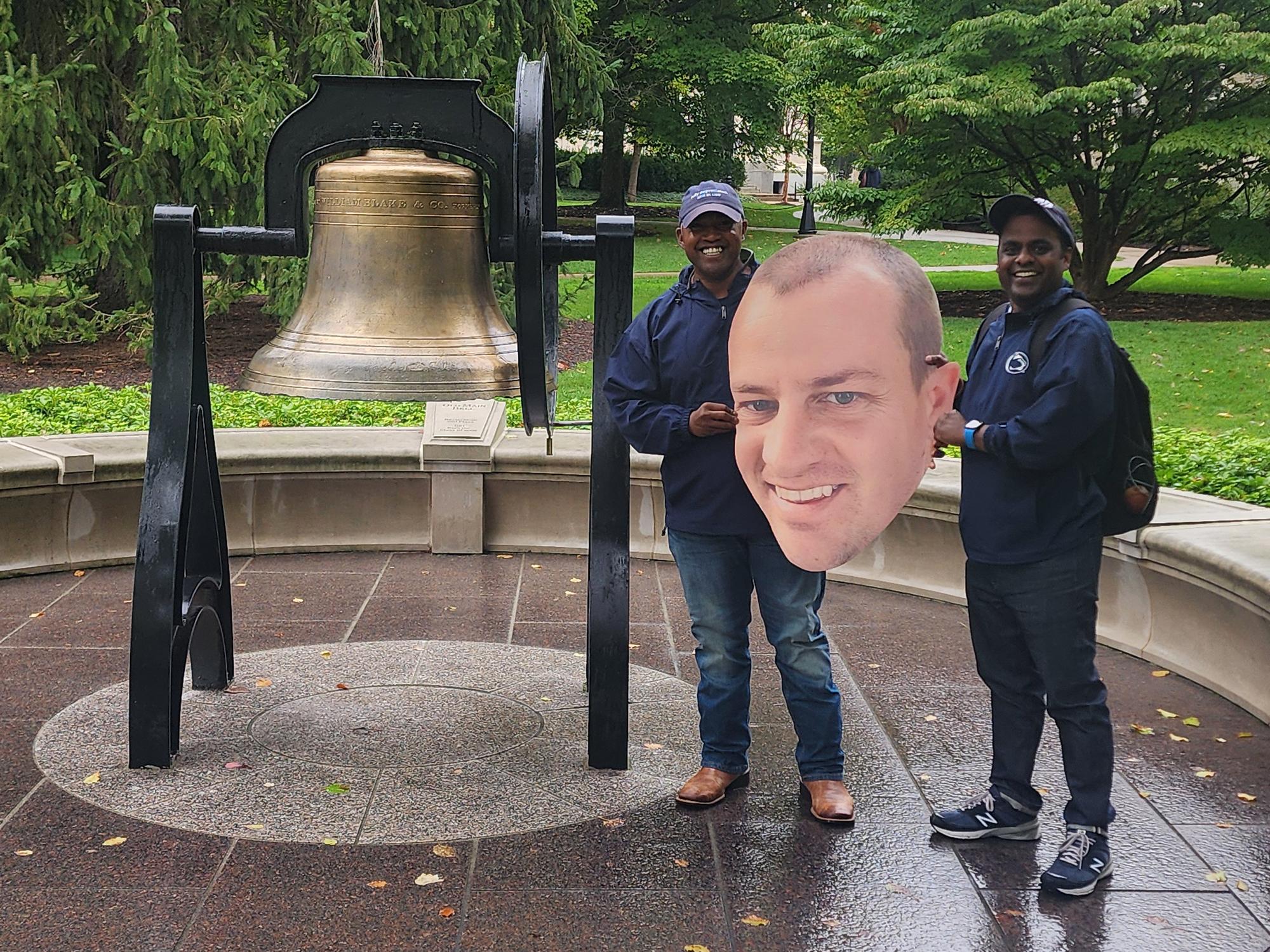 Two men pose in front of a bell with a printed cutout of a man's head.
