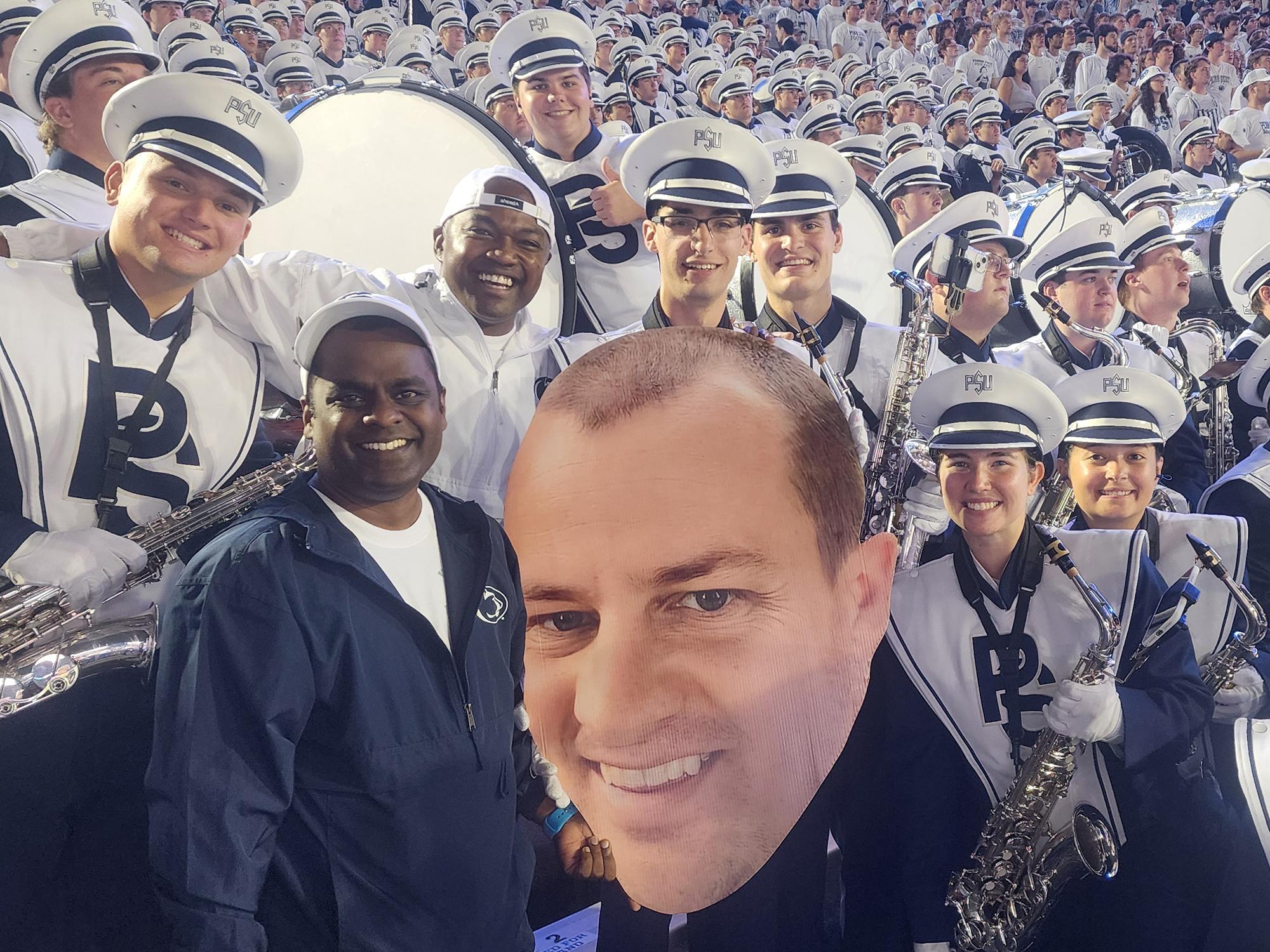 A group of people including the Penn State Blue Band pose with a large printed cutout of a man's face.