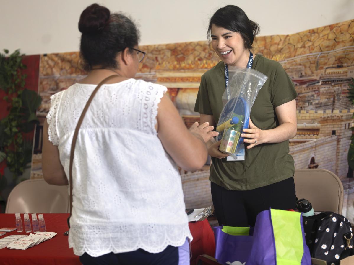 Two women face one another. The woman facing the camera is smiling and holding a plastic bag filled with medical supplies. The other woman faces her and gestures as the two talk. They are flanked by a table, upon which is a row of plastic bottles and pamphlets.