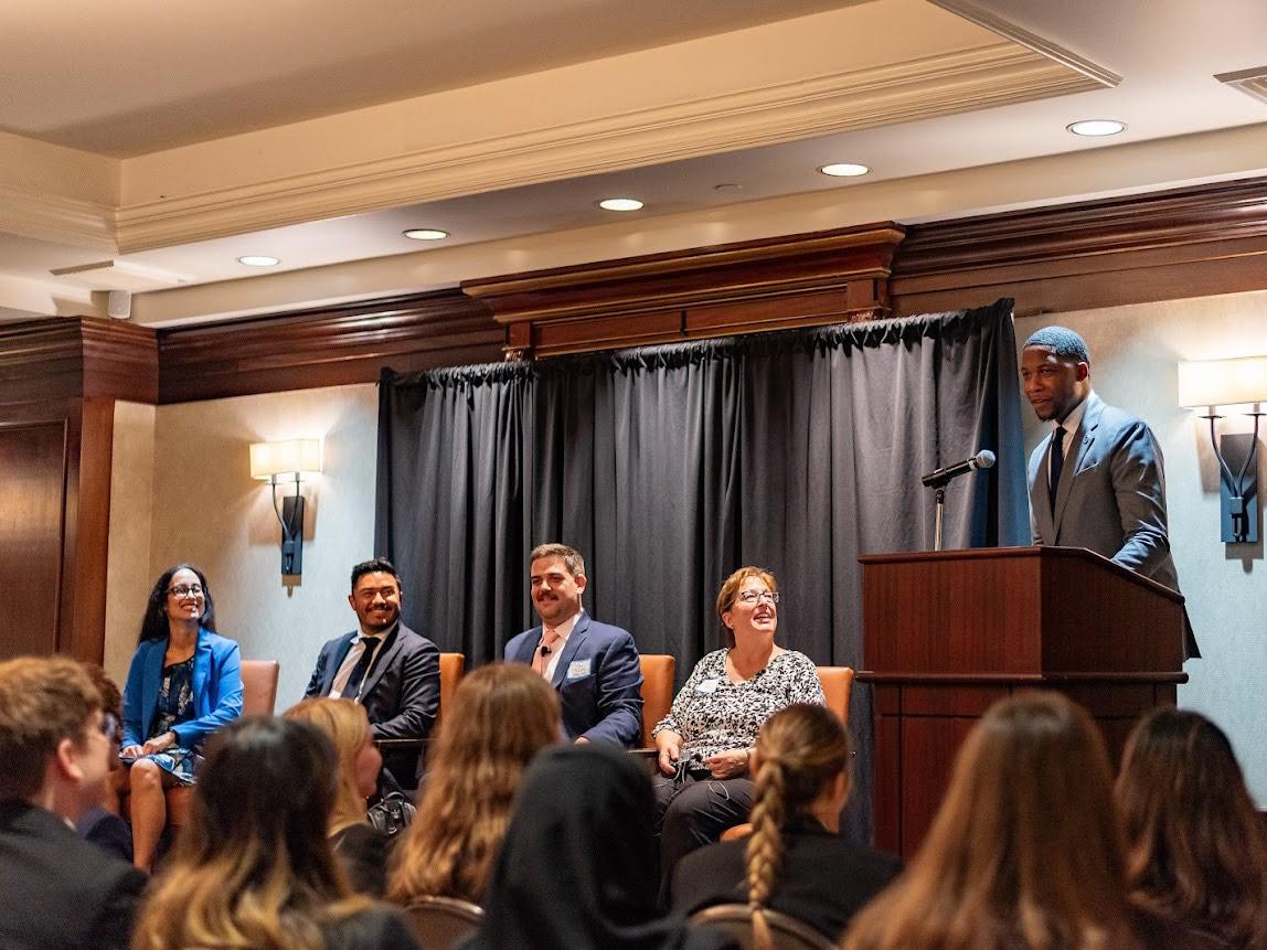 Anthony Mitchell stands at a podium leading his fellow alumni in a moderated conversation about careers in federal agencies and public service. Panelists from left to right: Evita Salles, Kevin Lima, Mike McCabe Jr. and Sharon Callahan.