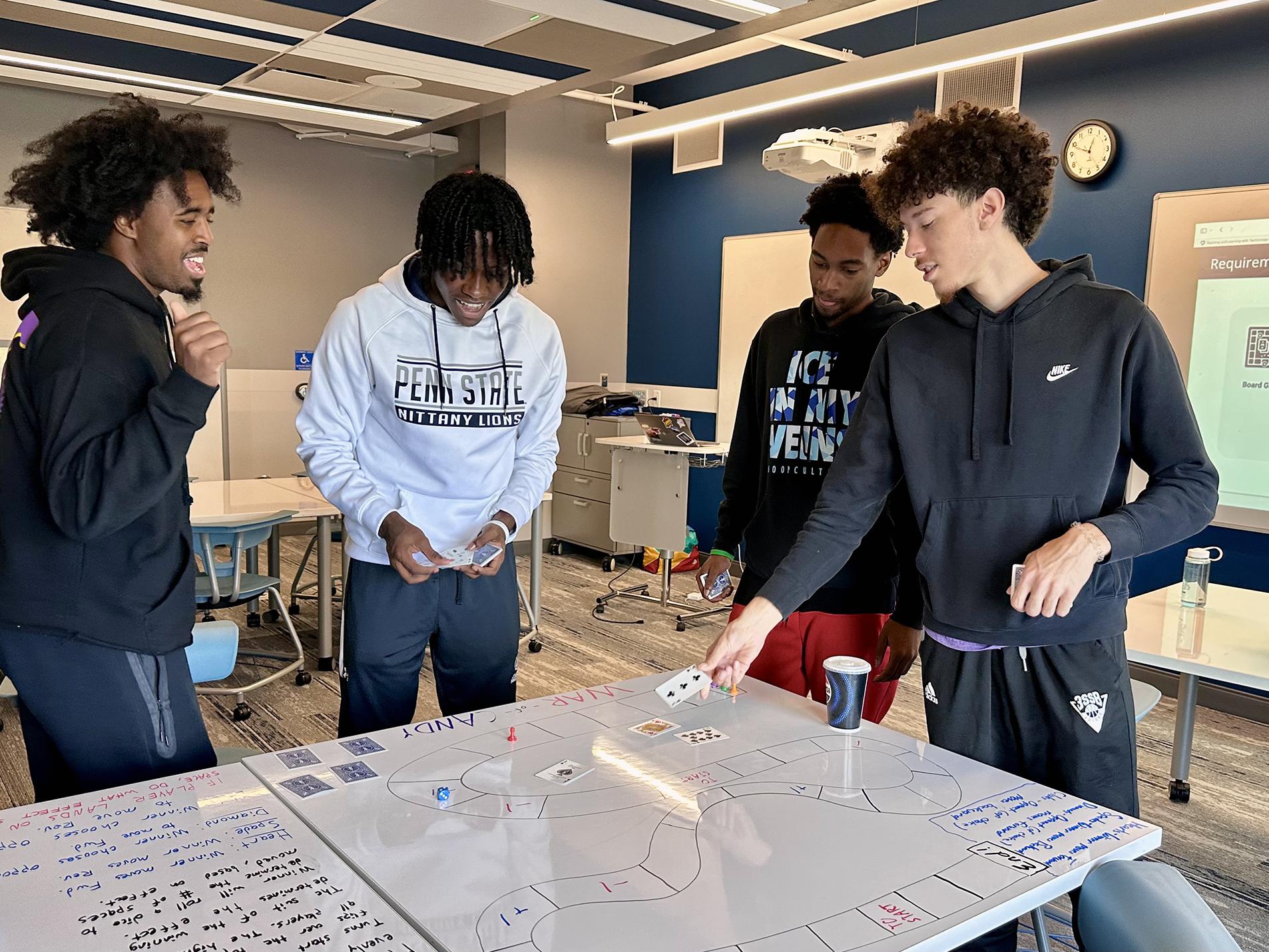 A group of four students stand around a whiteboard table with a custom-made board game on it