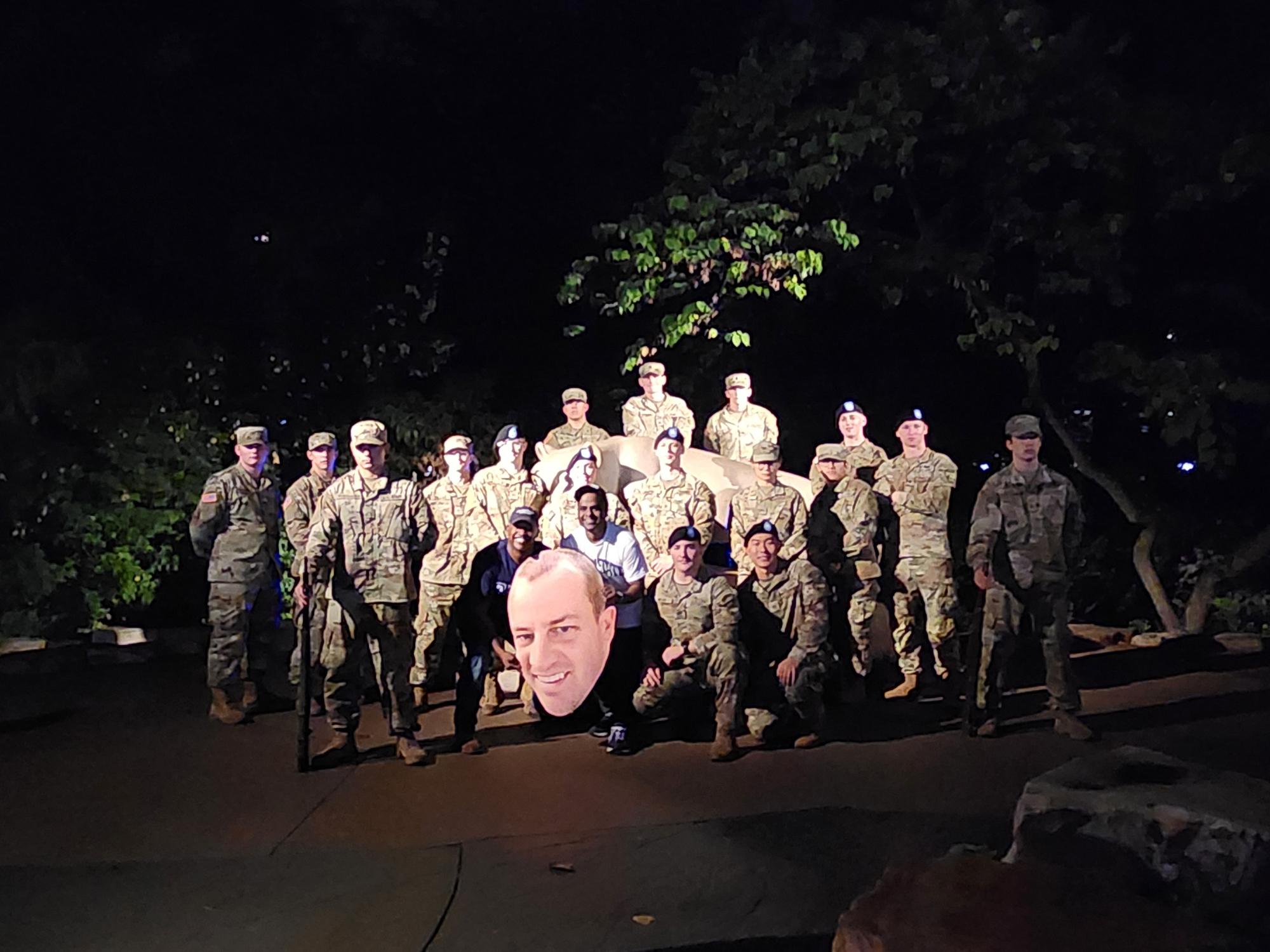 People in military uniforms stand around the Nittany Lion Shrine with a large printed cutout of a man's face.