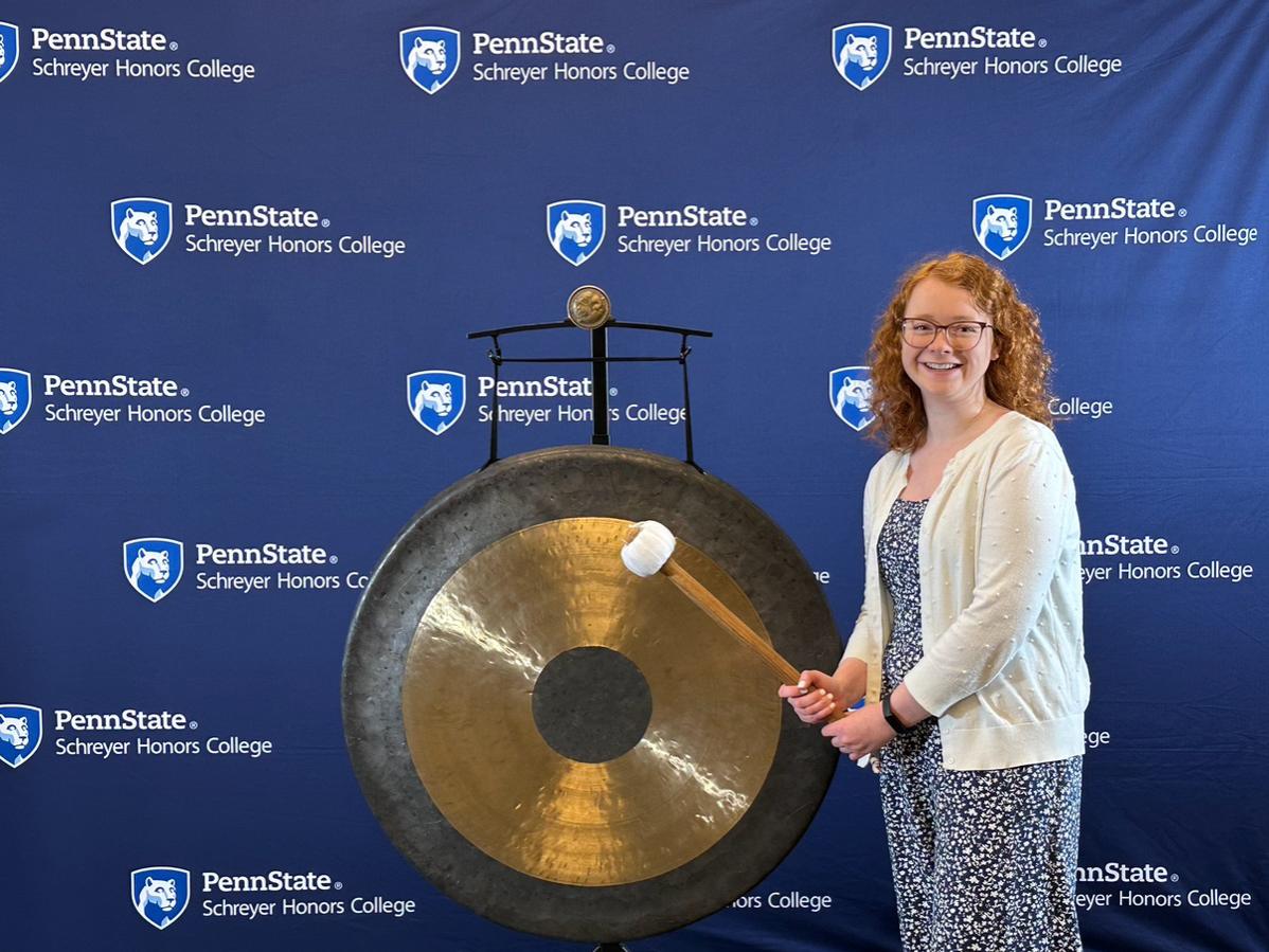 Julia Welp stands in front of a gong and a Penn State Schreyer Honors College backdrop to ring in her finishing her honors thesis.