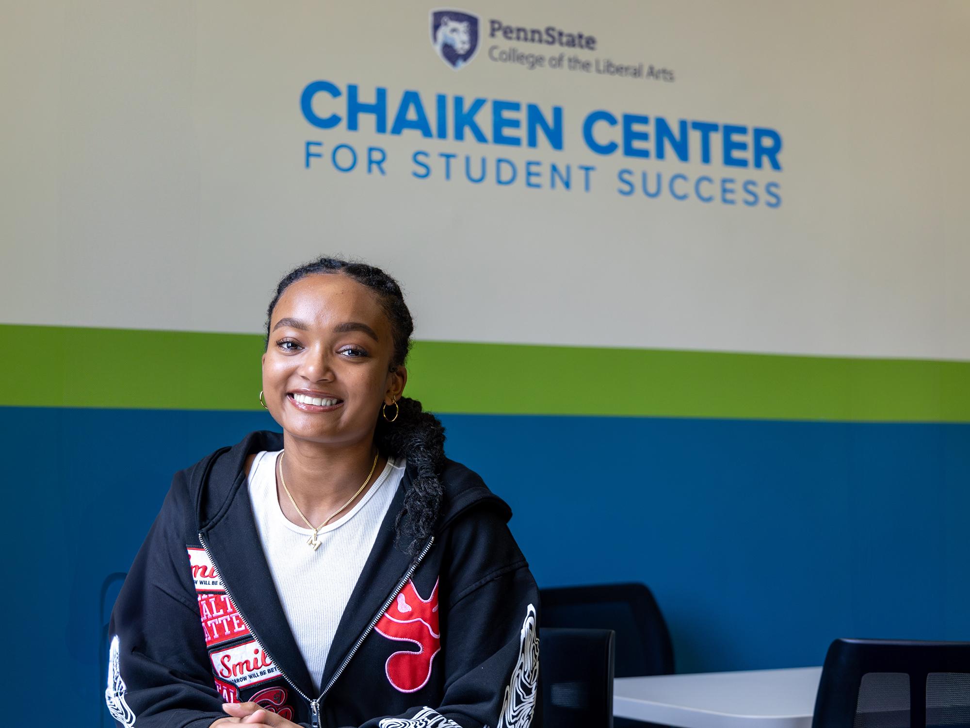 Milan Dukes sits in the Liberal Arts Chaiken Center for Student Success with the office’s word mark and the Penn State Liberal Arts word mark on the wall behind her.