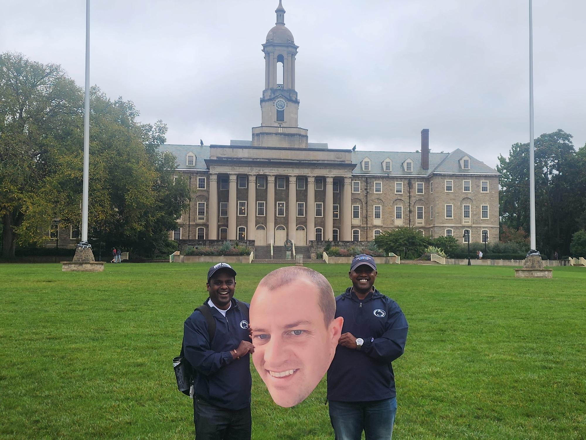 Two men pose in front of Old Main with a printed cutout of a man's head.