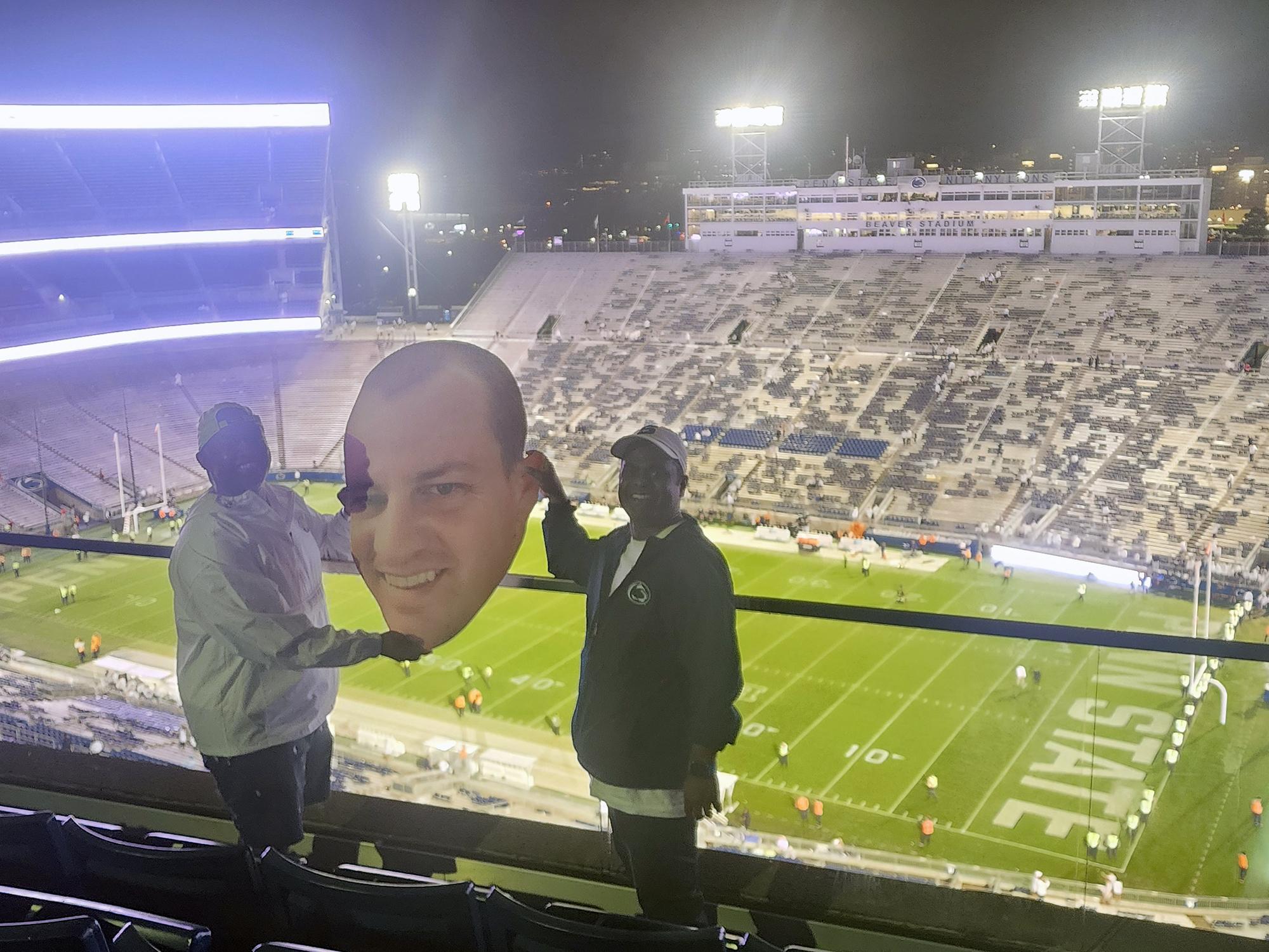 Two men pose inside Beaver Stadium with a printed cutout of a man's head.