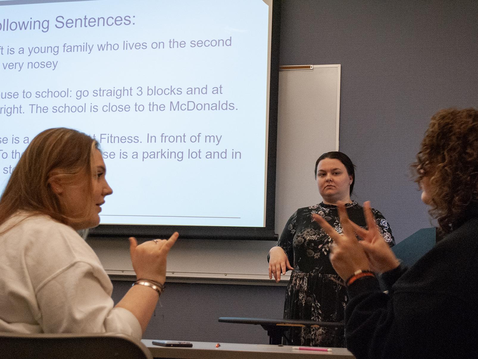 Two students face each other and use sign language, while an instructor watches