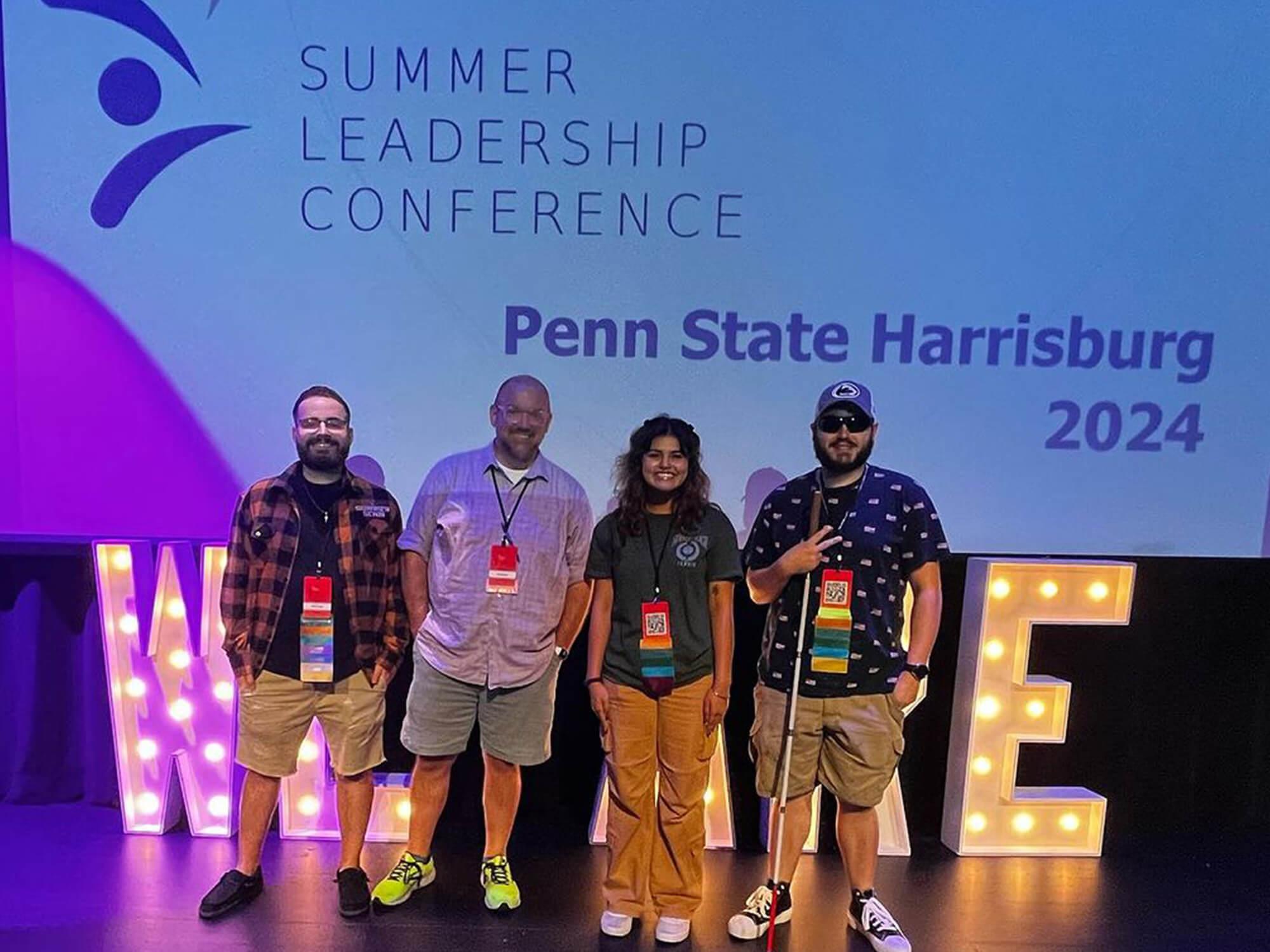 A group of four individuals poses in front of a projector screen that reads “Summer Leadership Conference” and “Penn State Harrisburg 2024.”