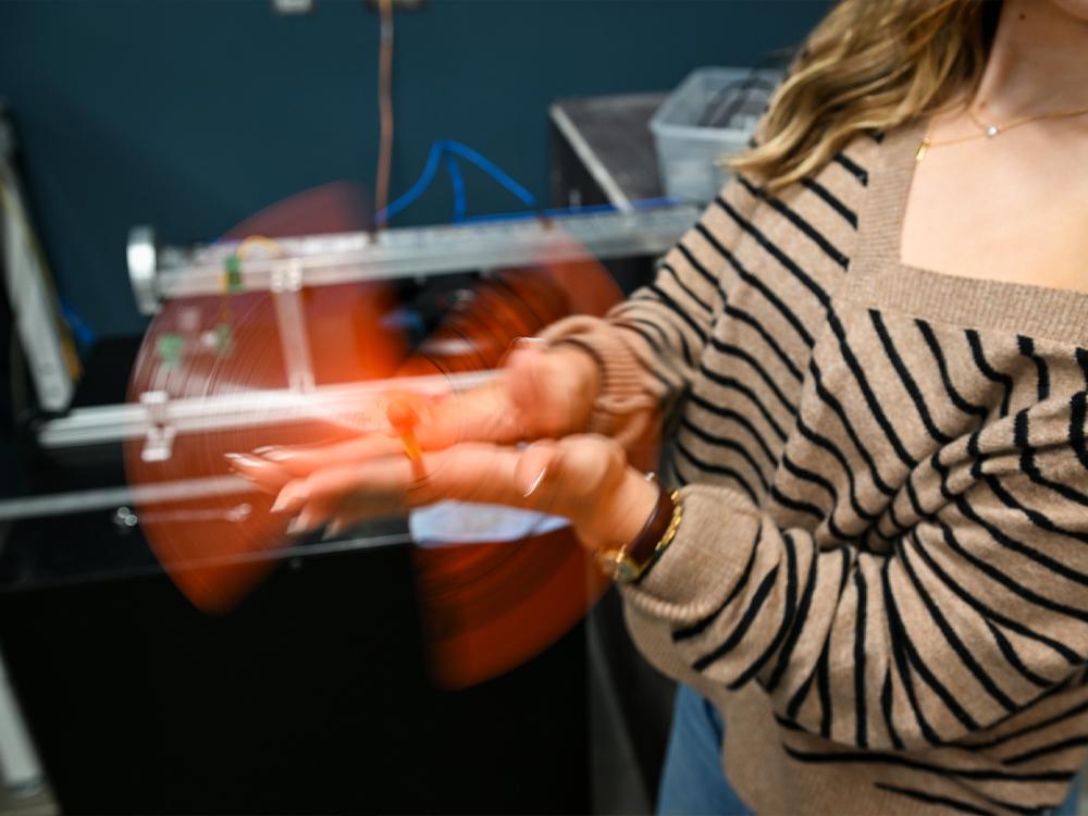 An individual in a striped, tan sweater spins an orange spinner toy in her hands in a lab setting.  