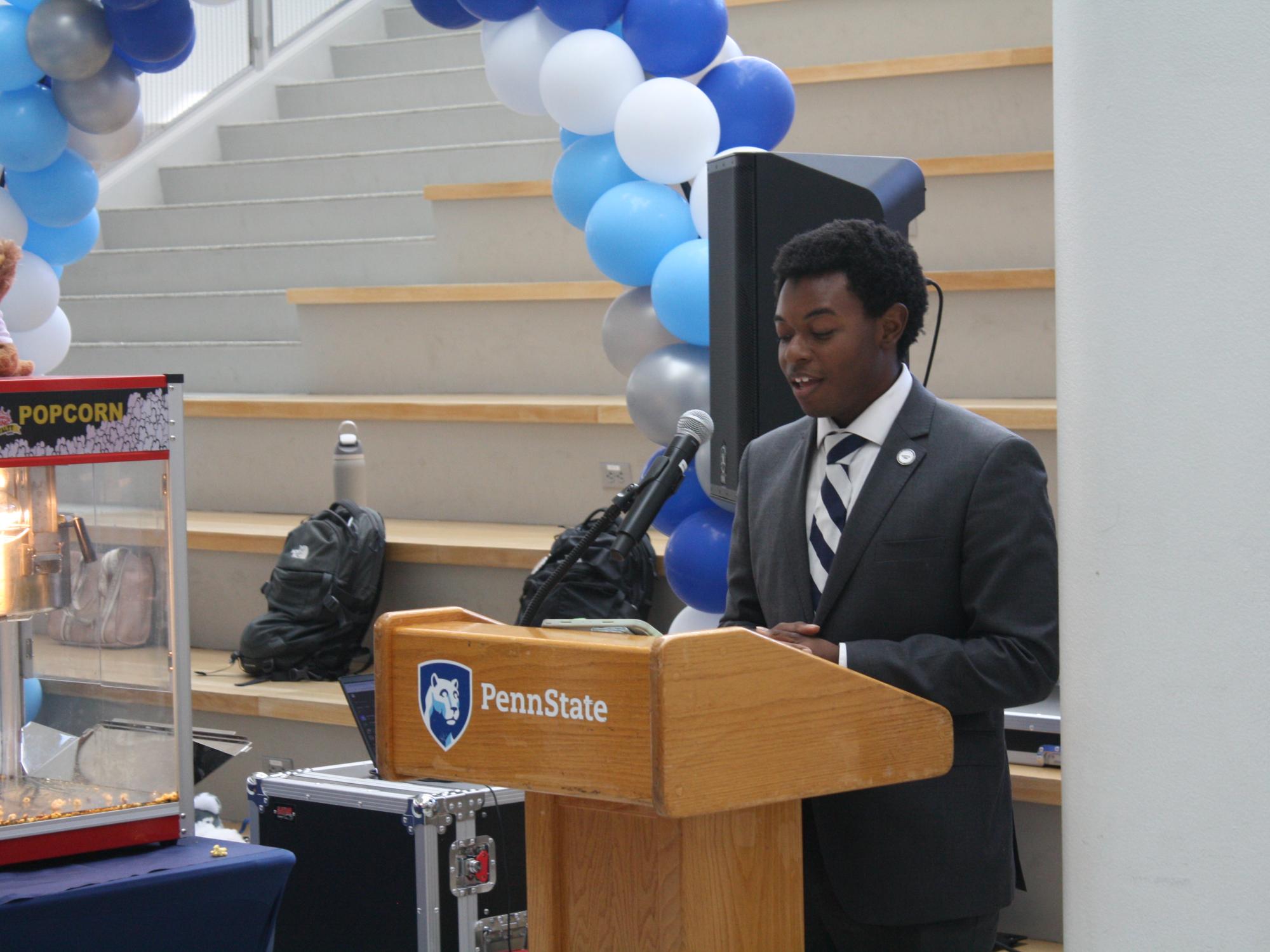 Zion Sykes speaks at a podium with a blue and white balloon arch in the background