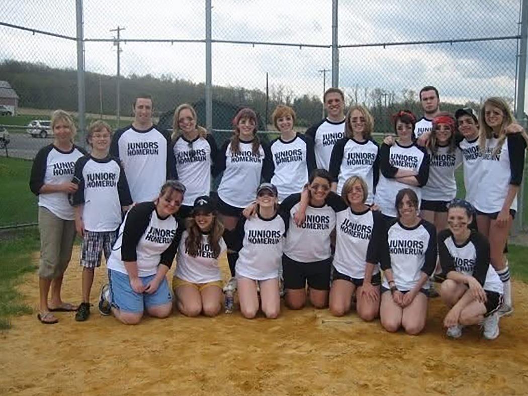 A group of students and teachers pose by the backstop on a softball field in shirts that read Juniors Homerun. 