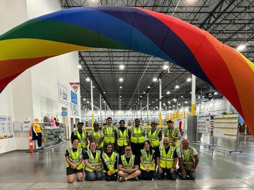 Two rows of Amazon works swearing safety vests while smiling for a picture under an inflatable rainbow in an Amazon warehouse.