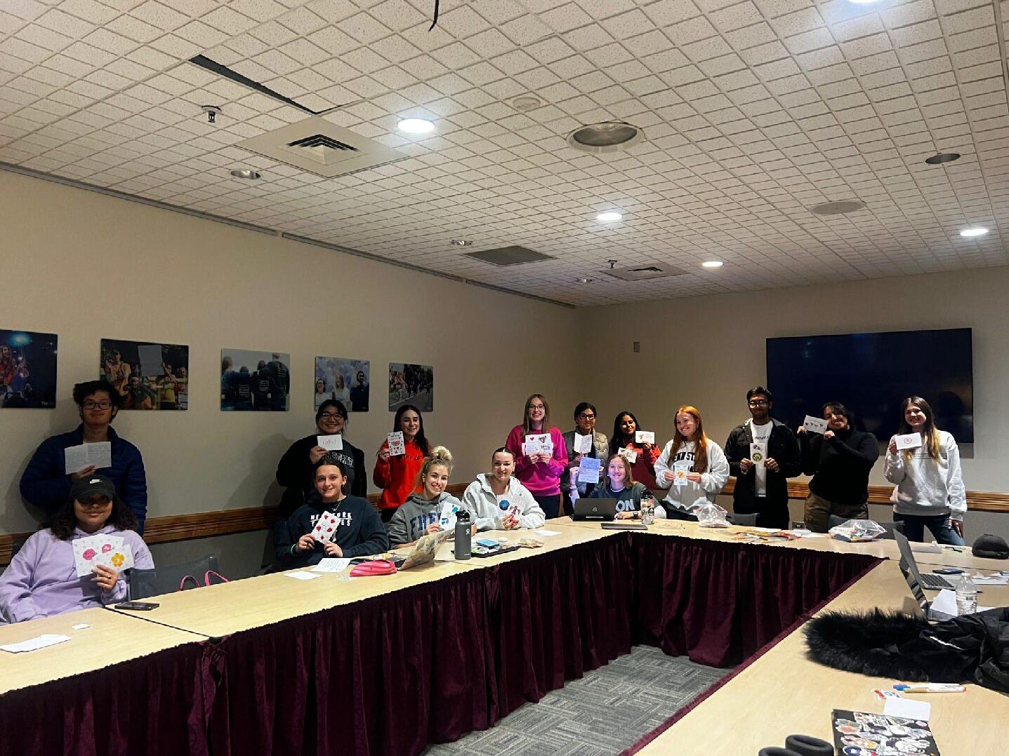 A group of students sit and stand around a conference table in a meeting room while holding up greeting cards.