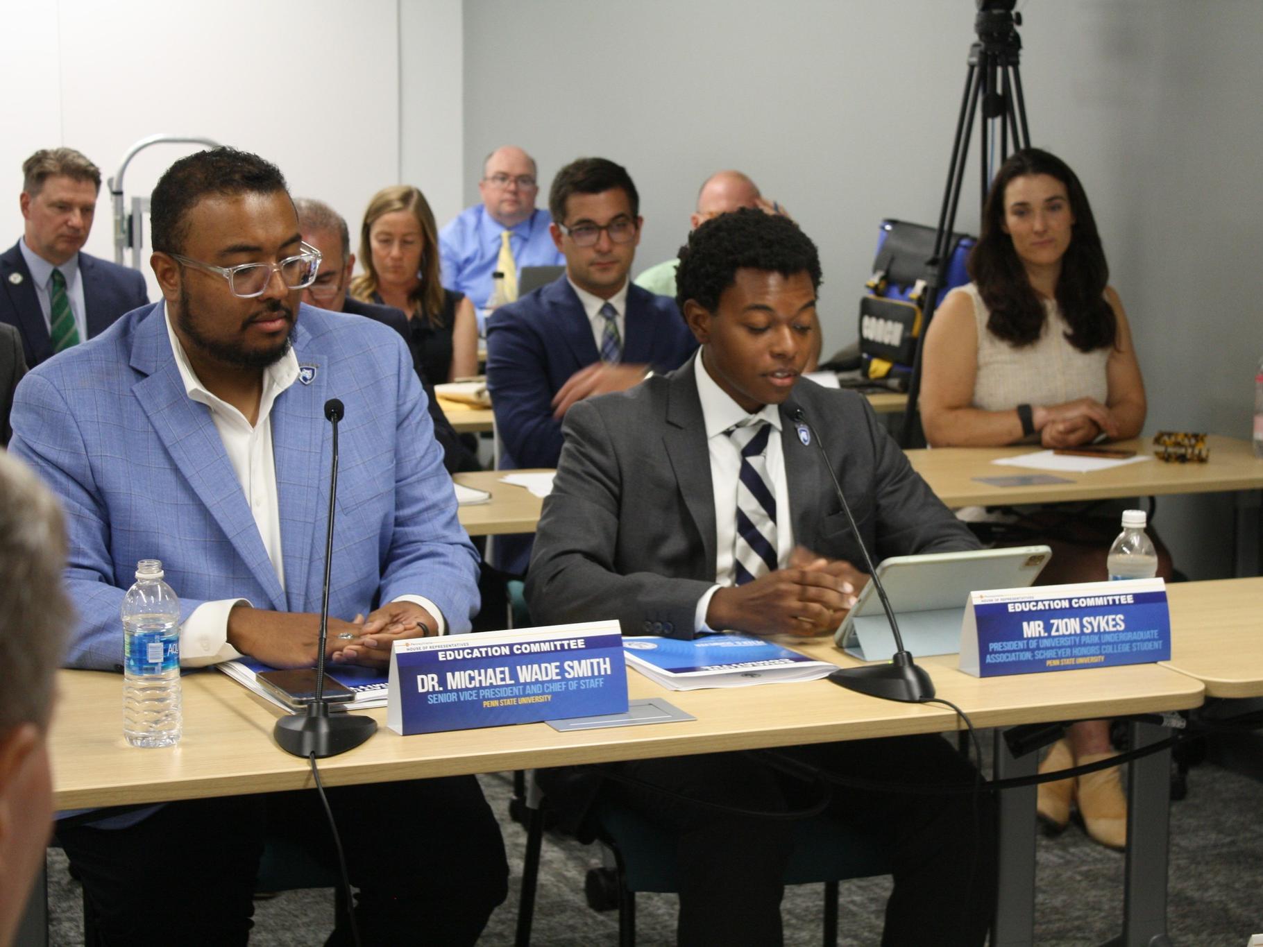 Two individuals wearing suits sit at a panel table with microphones in front of them