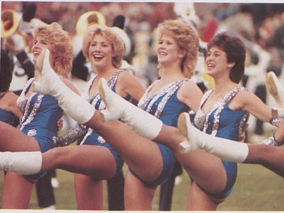 Majorettes wearing blue bodysuits kick their legs in the air on the football field in the 1980s