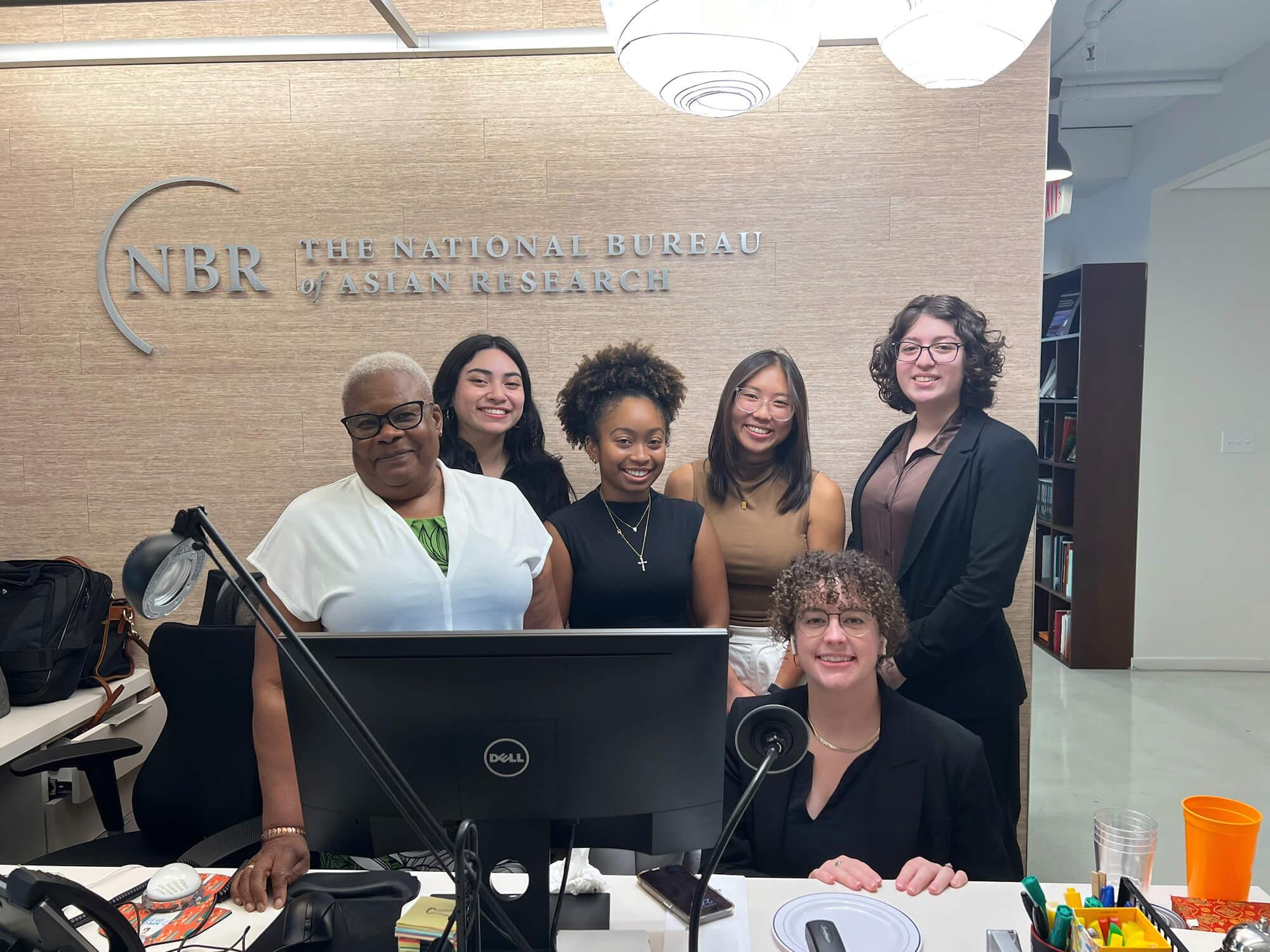 Charis Liu (back row, second from right) stands with five other people behind a desk and in front of a sign on the wall that reads “NBR The National Bureau of Asian Research.”
