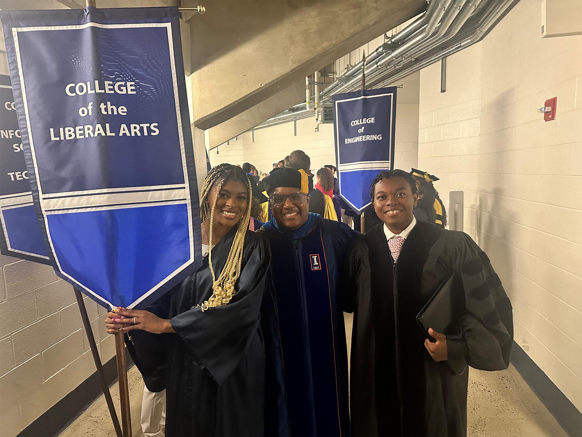 Zion Sykes (right) — pictured with Alana Nesmith (left) and Clarence Lang, Susan Welch Dean of the College of the Liberal Arts (middle) — wear commencement robes and hold a banner that says “College of the Liberal Arts” in a back hallway of the Bryce Jordan Center. 