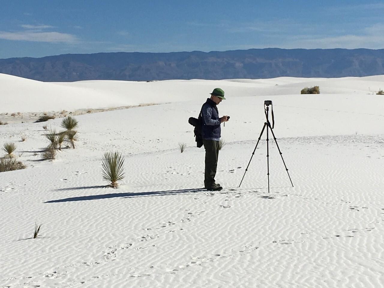 A man stands in front of a camera on a tripod in the middle of a desert. 