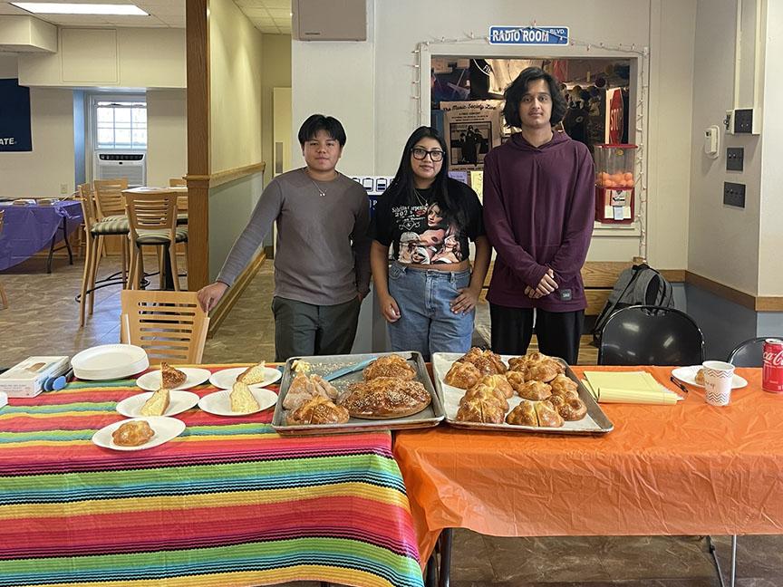 Three people standing behind a table with food placed on a colorful tablecloth.