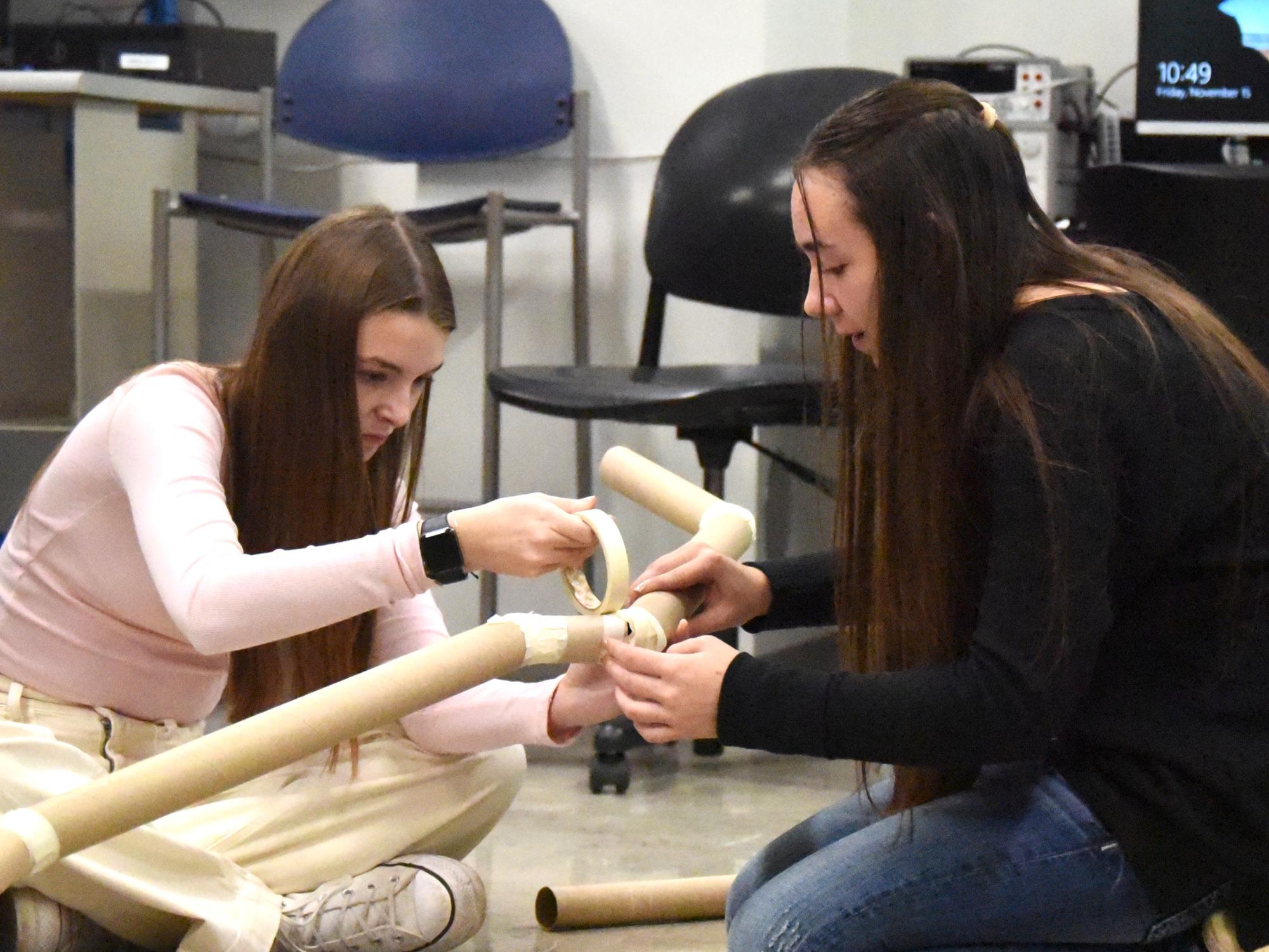 Two female high-school students build a model pipeline with cardboard tubes and masking tape.