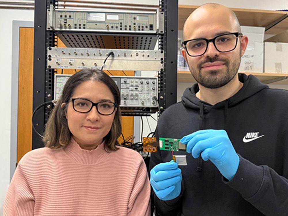 Two people wearing glasses pose in a lab. One holds an electronic sensing device.