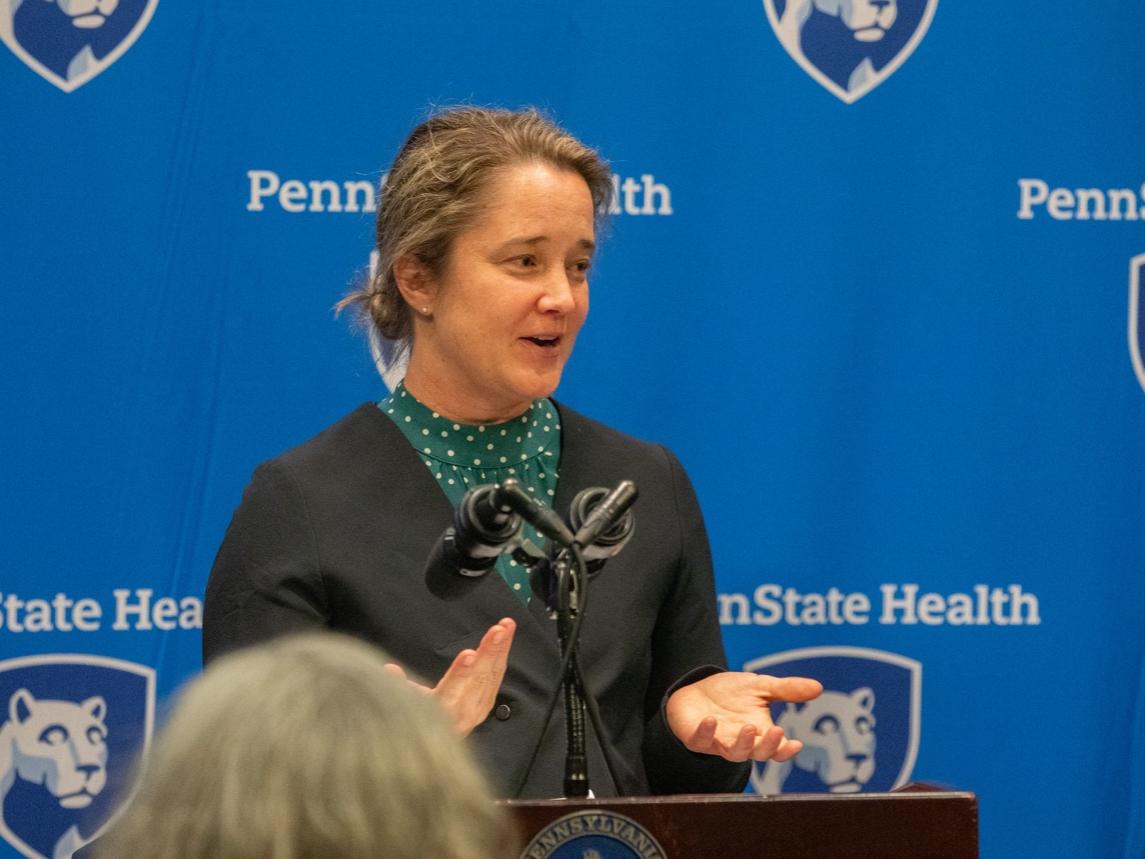 A woman in professional attire speaks at a lectern, gesturing with her hands. A step-and-repeat ‘Penn State Health’ backdrop is in the background.