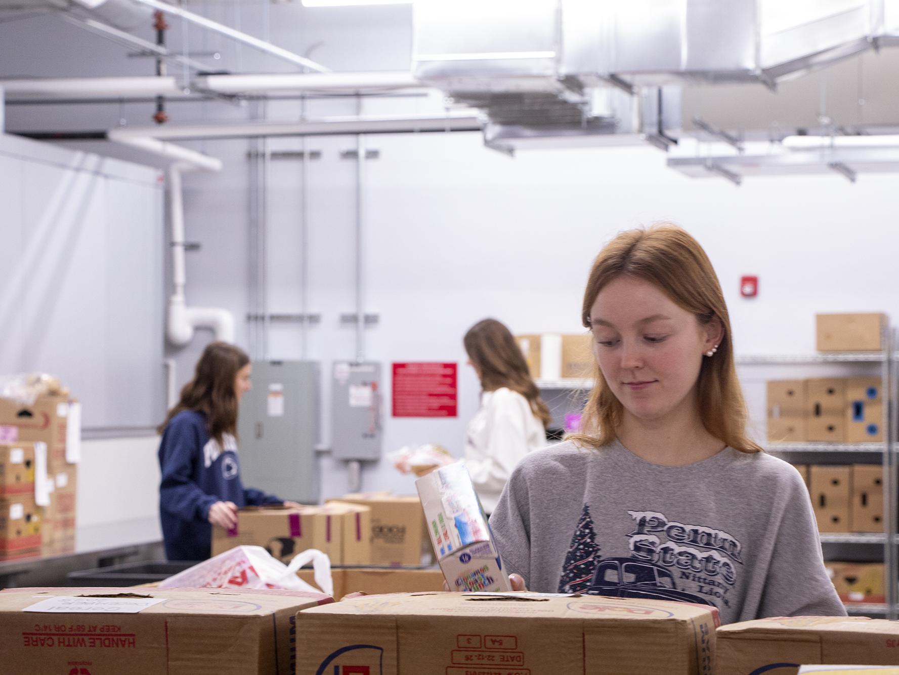 A student organizing a box of donated food items at the State College Food Bank, while other students similarly work behind her.