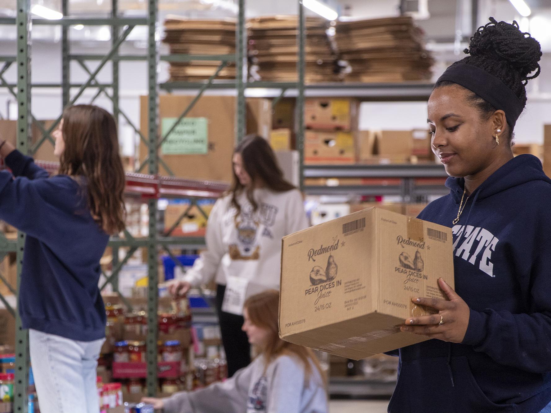 A student carrying a box at the State College Food Bank, while other students behind her organize shelves.