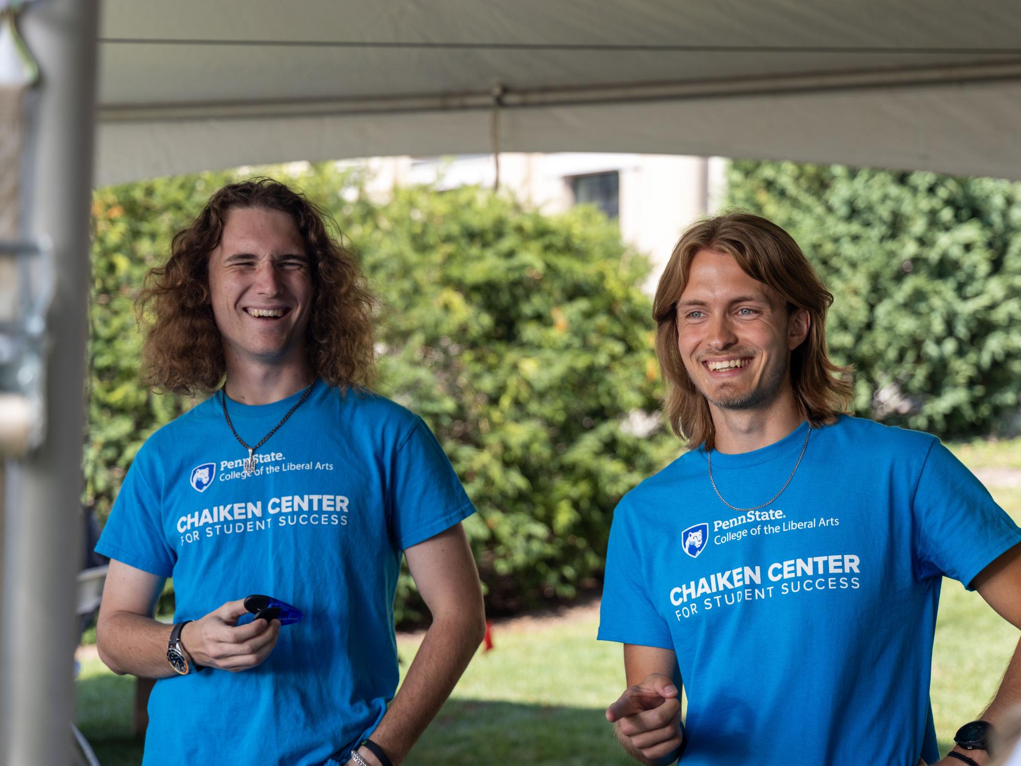 Igor Latsanych (left) and Victor Frolenko (right) laugh while wearing blue shirts with the Penn State Liberal Arts wordmark and “Chaiken Center for Student Success” on them while standing under a tent on Pattee Mall.