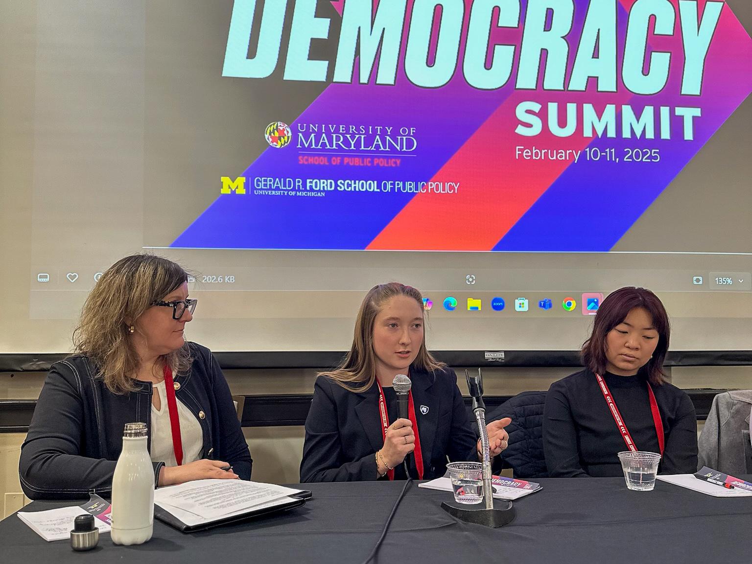 Madelyn Hindman holds a microphone while speaking on a panel. She sits between two other speakers.