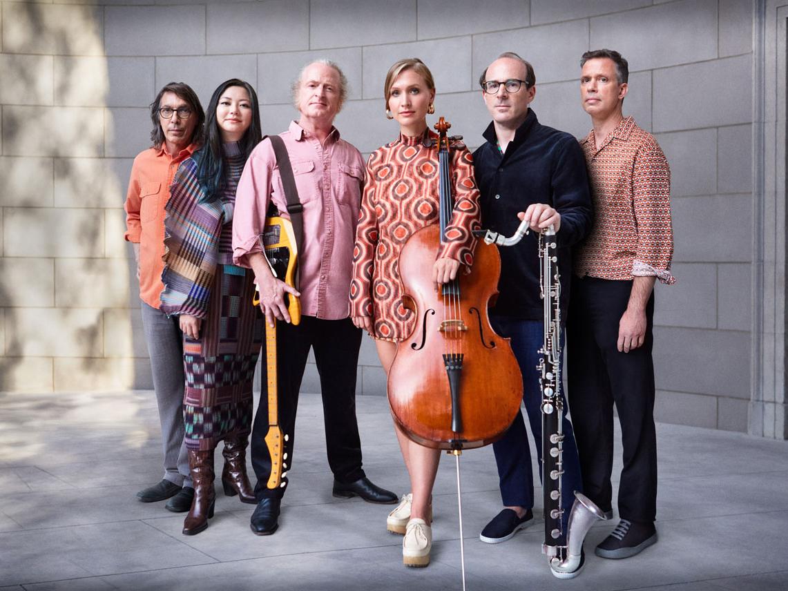 A diverse group of six musicians stand near a wall.