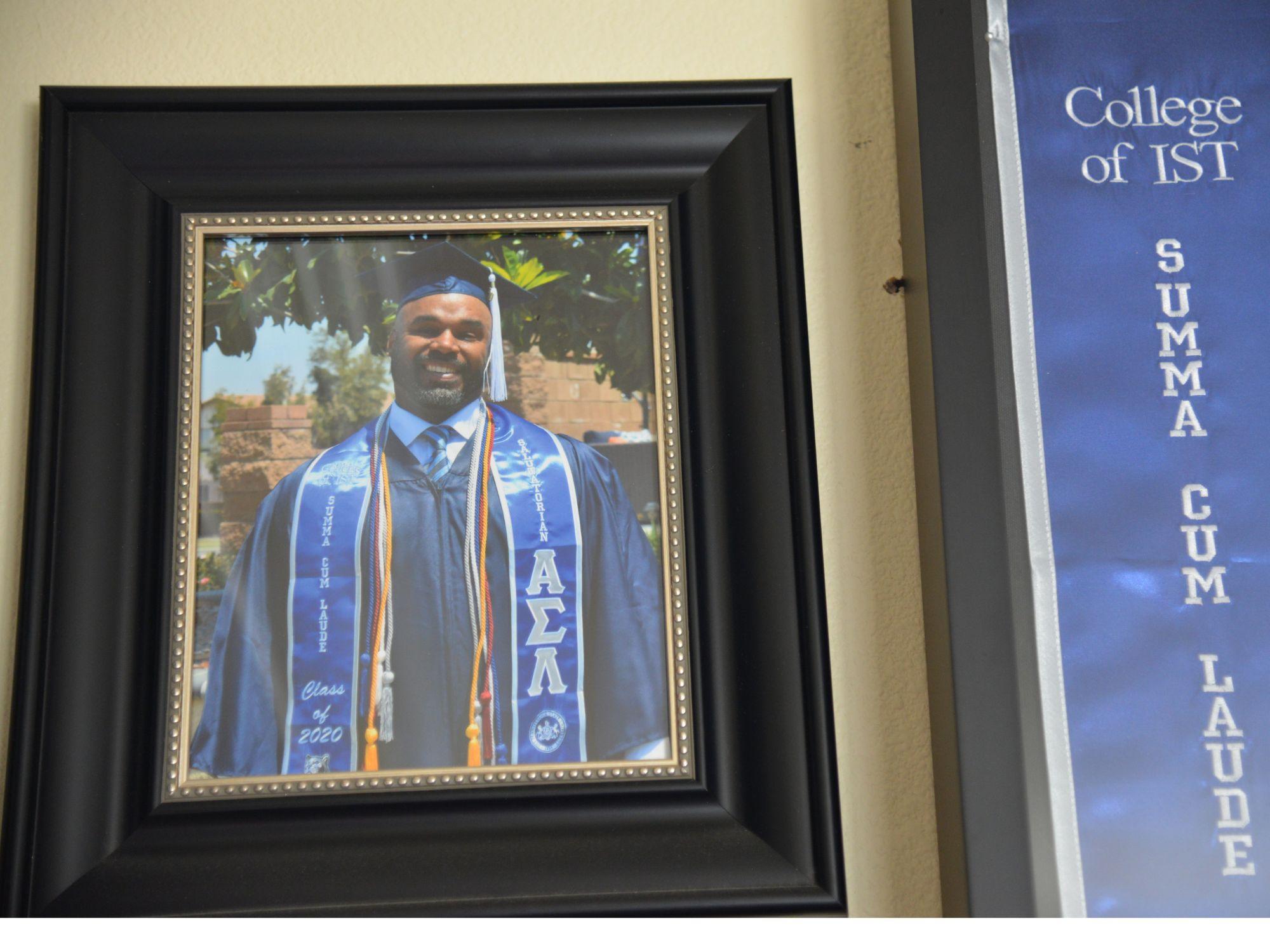 A picture framed of corey Wheeler wearing his graduation cap and gown is next to a framed banner from the College of IST