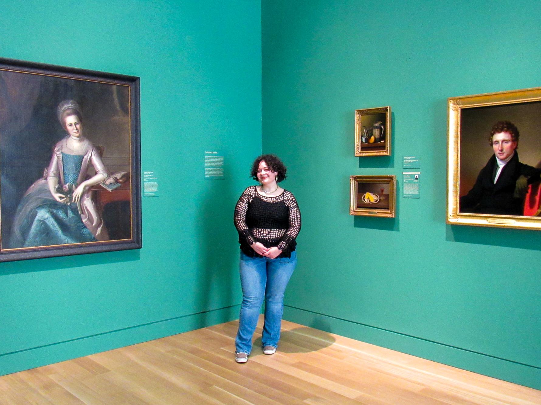 Amanda Demuth stands in front of two sea foam green walls among four paintings in the Palmer Museum of Art.