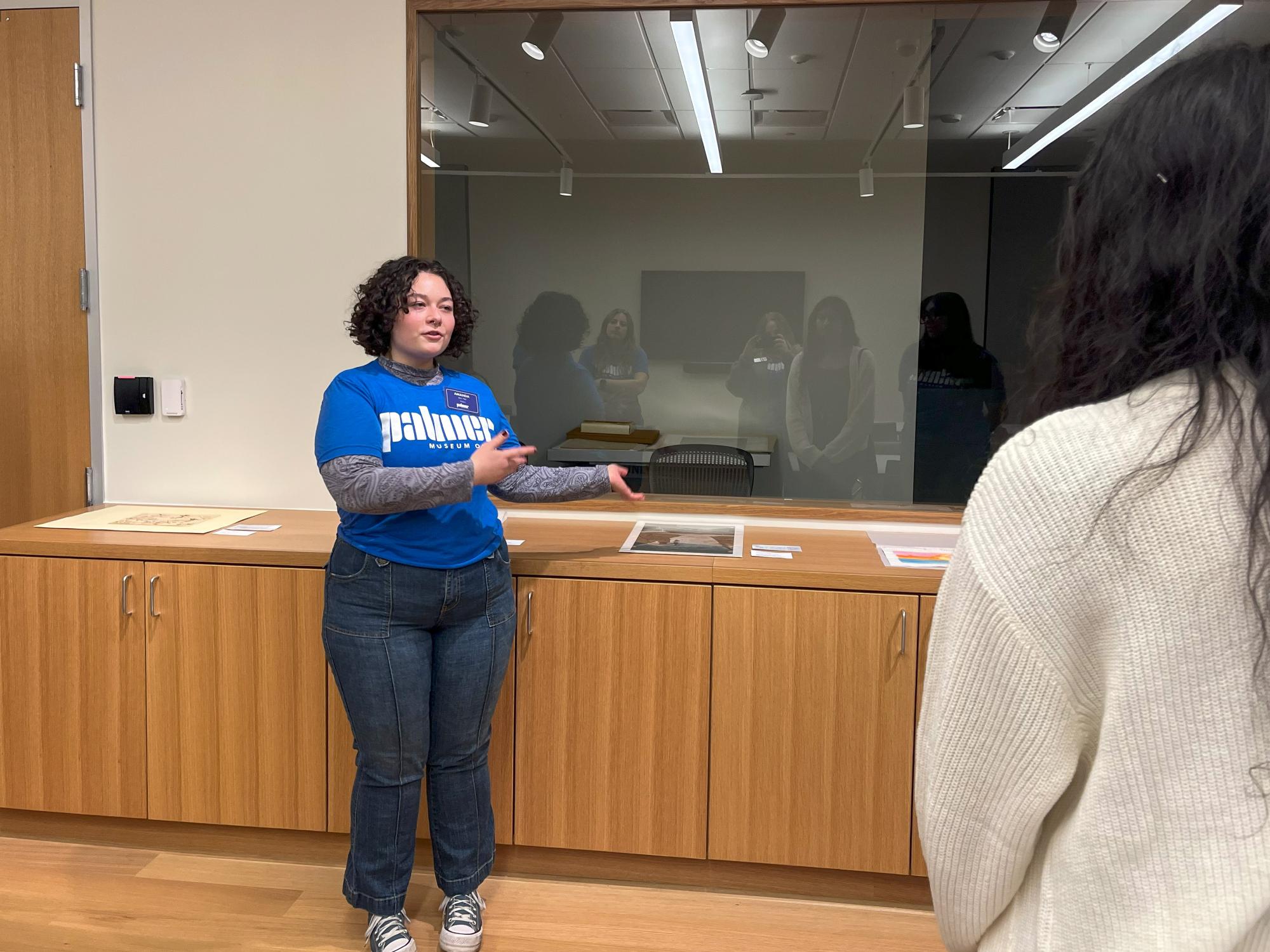 Amanda Demuth presents at the front of a classroom in the Palmer Museum of Art while wearing a blue shirt that reads “Palmer Museum of Art.”