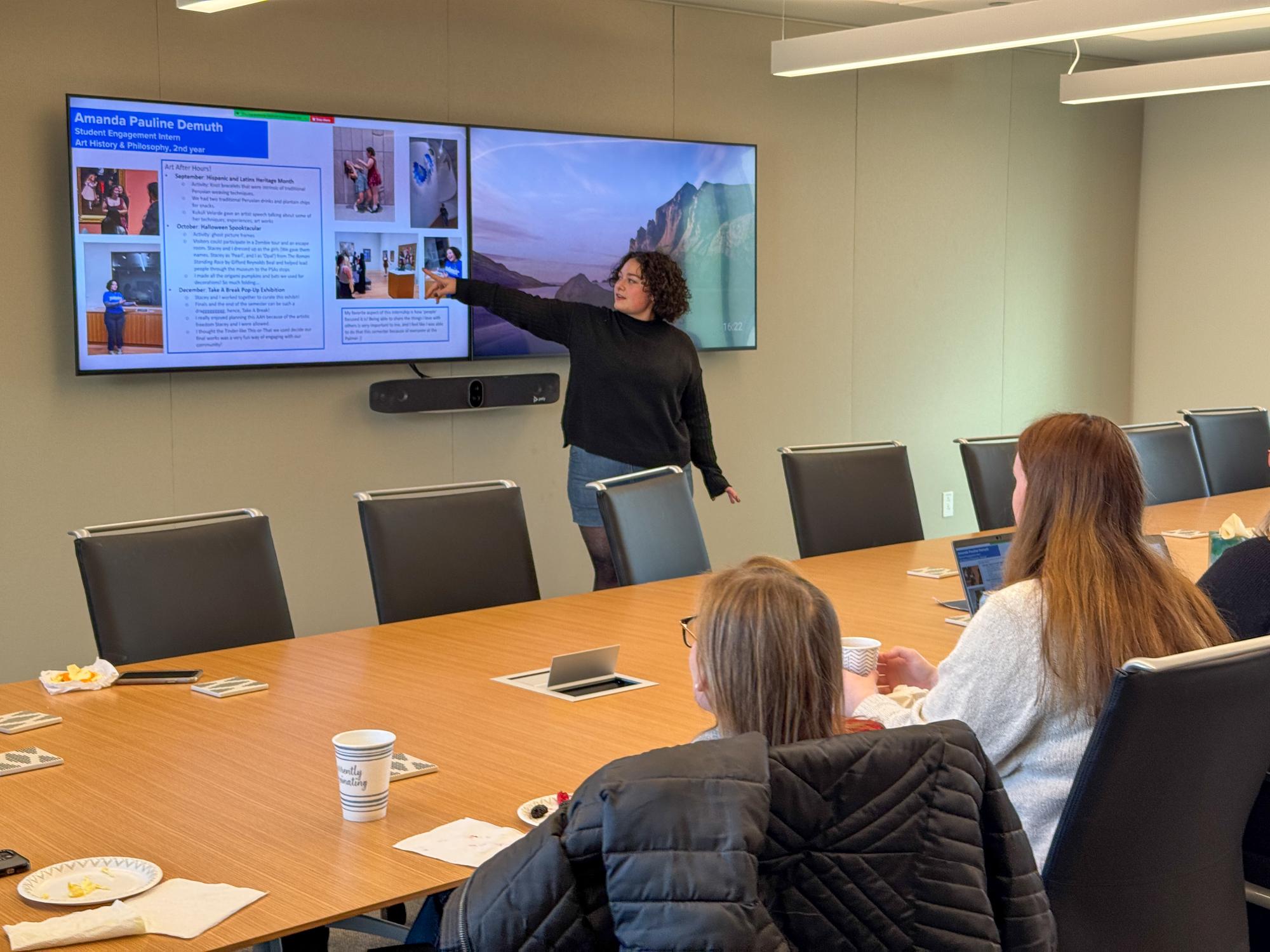 Amanda Demuth points at a TV screen with text and photos about herself while standing at the front of a conference room.