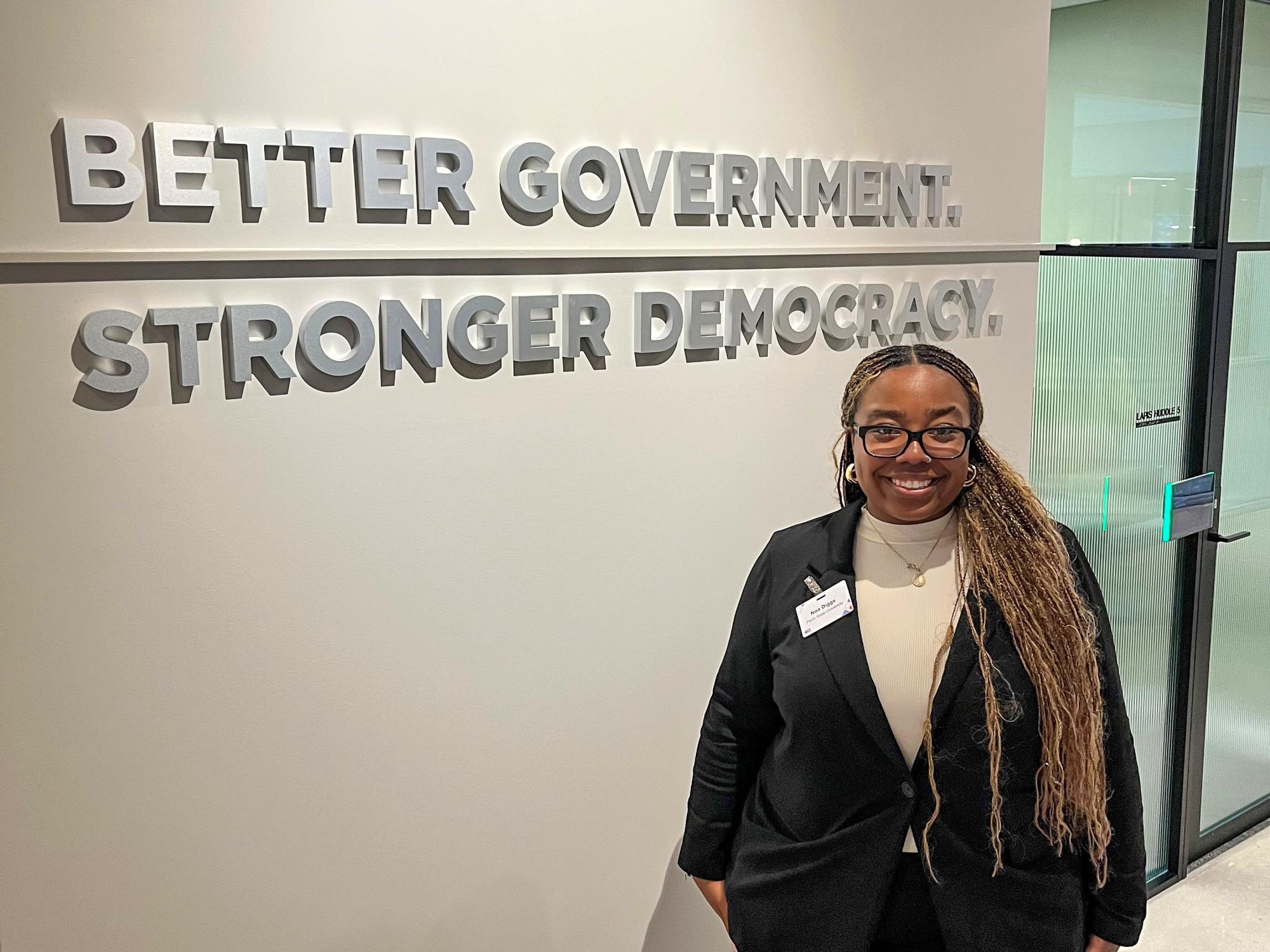 Noa Diggs stands in front of a sign on a wall that reads “Better Government. Stronger Democracy.”