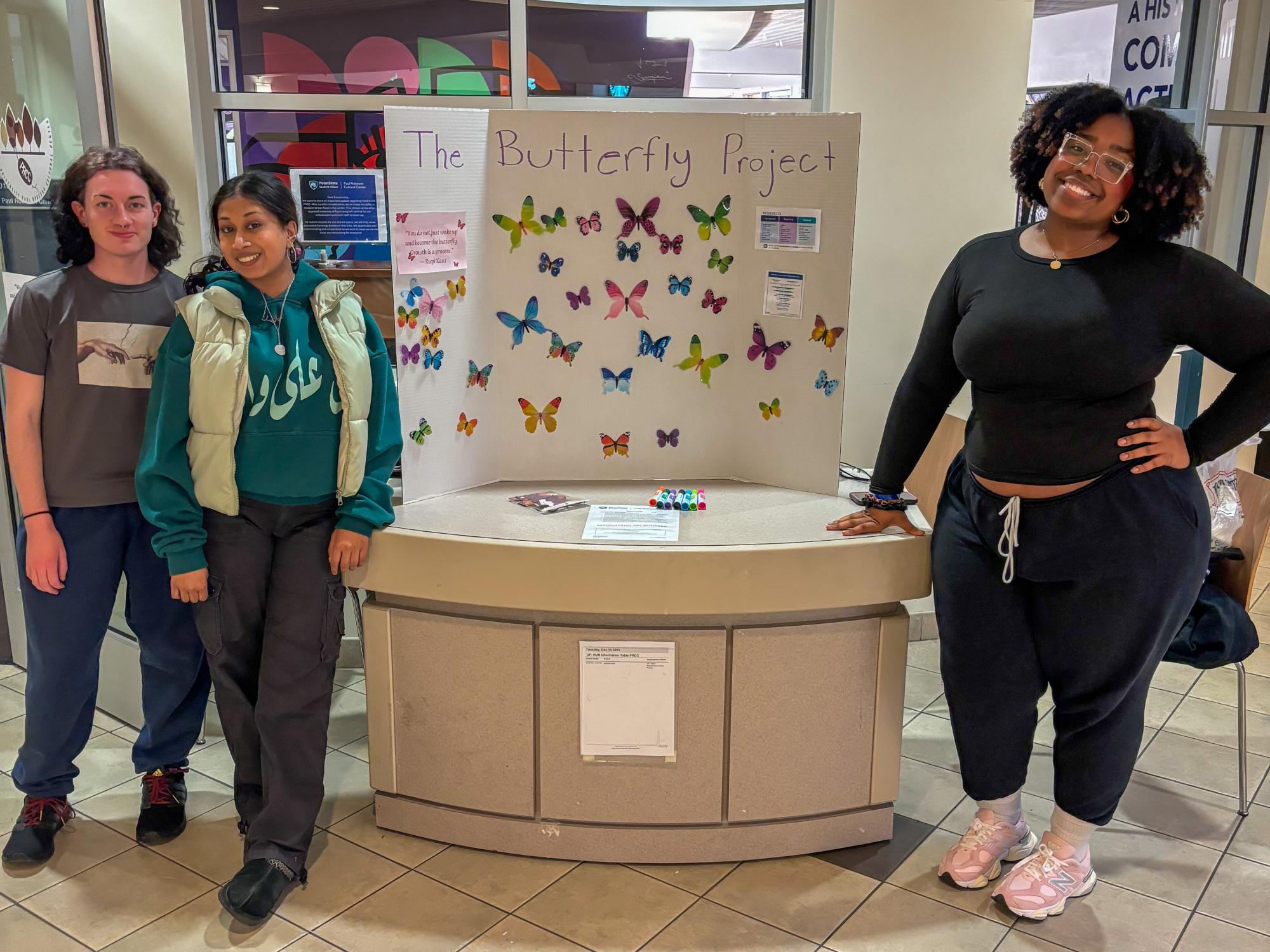 Noa Diggs (right) stands to the right and two other students stand to the left of a poster board that reads “The Butterfly Project” and has photos of butterflies on it.